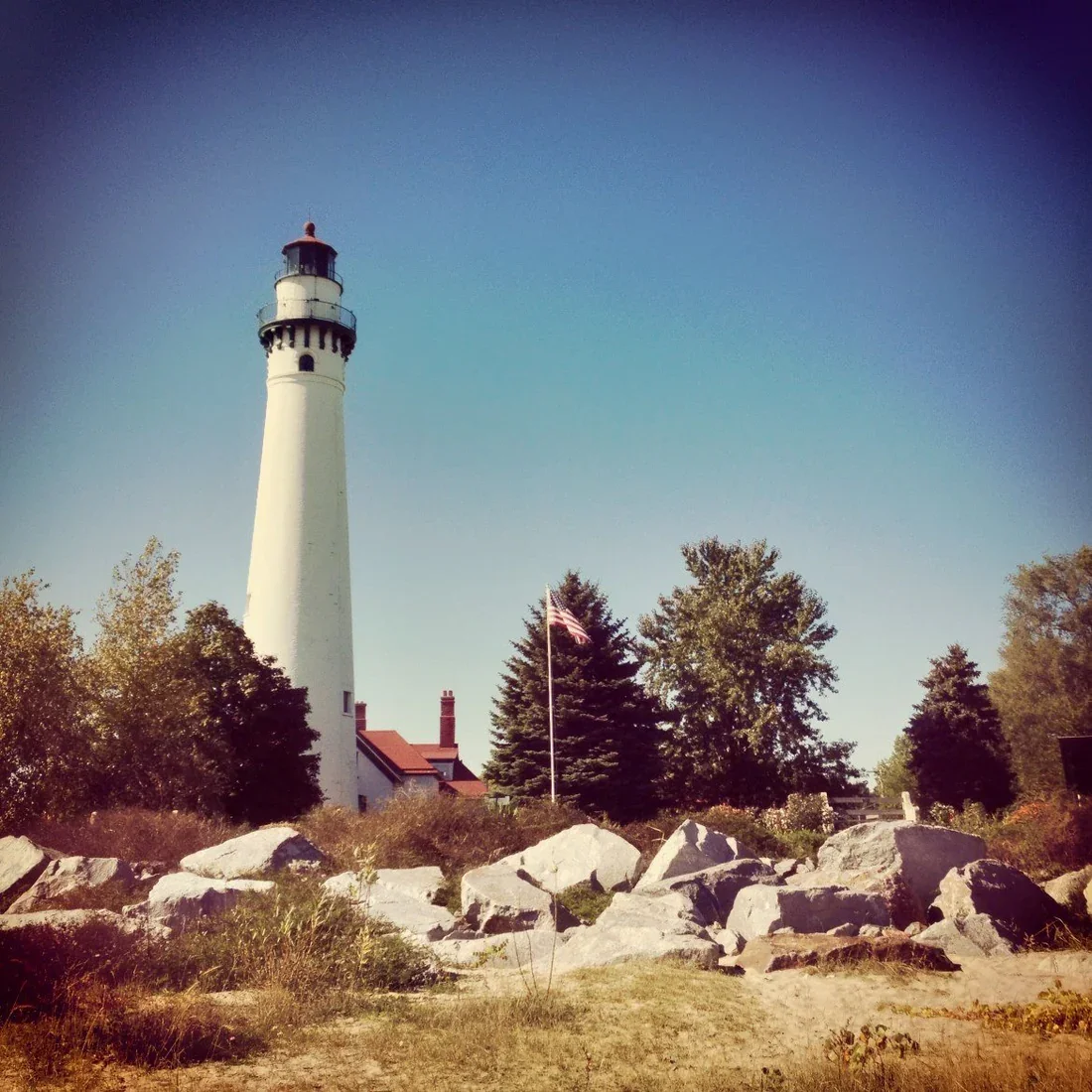 A tall white lighthouse with a red top stands near trees and rocks, with an American flag flying on a flagpole in front. Clear blue sky in the background.