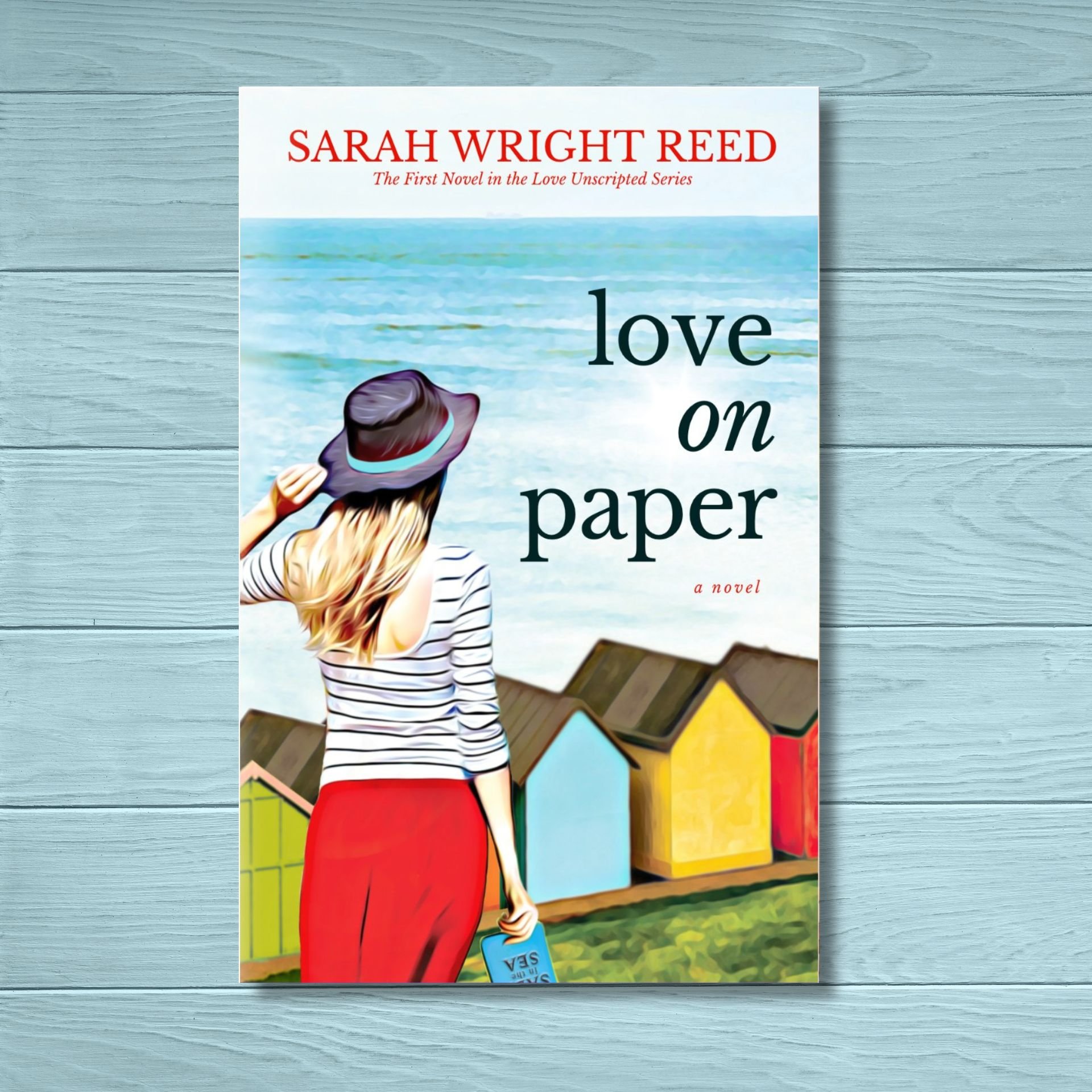 Book cover titled 'Love on Paper' by Sarah Wright Reed, featuring a woman with long blonde hair wearing a striped shirt and red pants, holding a blue book with a background of colorful houses and a blue sky.