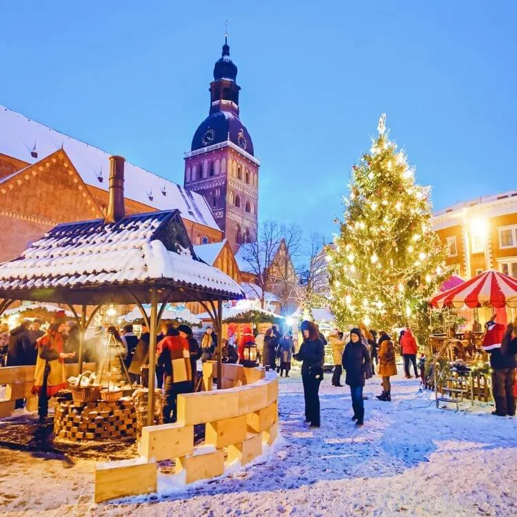 A snowy Christmas market scene in a European town square with a decorated Christmas tree, festive stalls, and a church tower in the background during evening.
