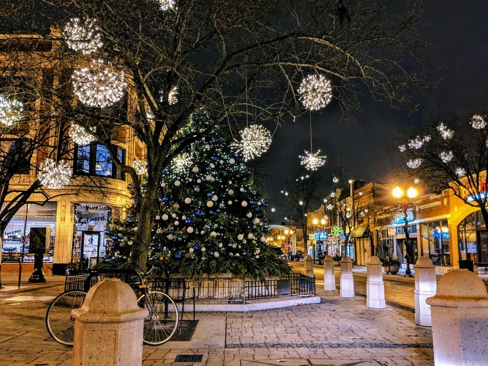 A decorated Christmas tree in an outdoor town square at night, with string lights and illuminated ornaments, surrounded by storefronts and street lamps.