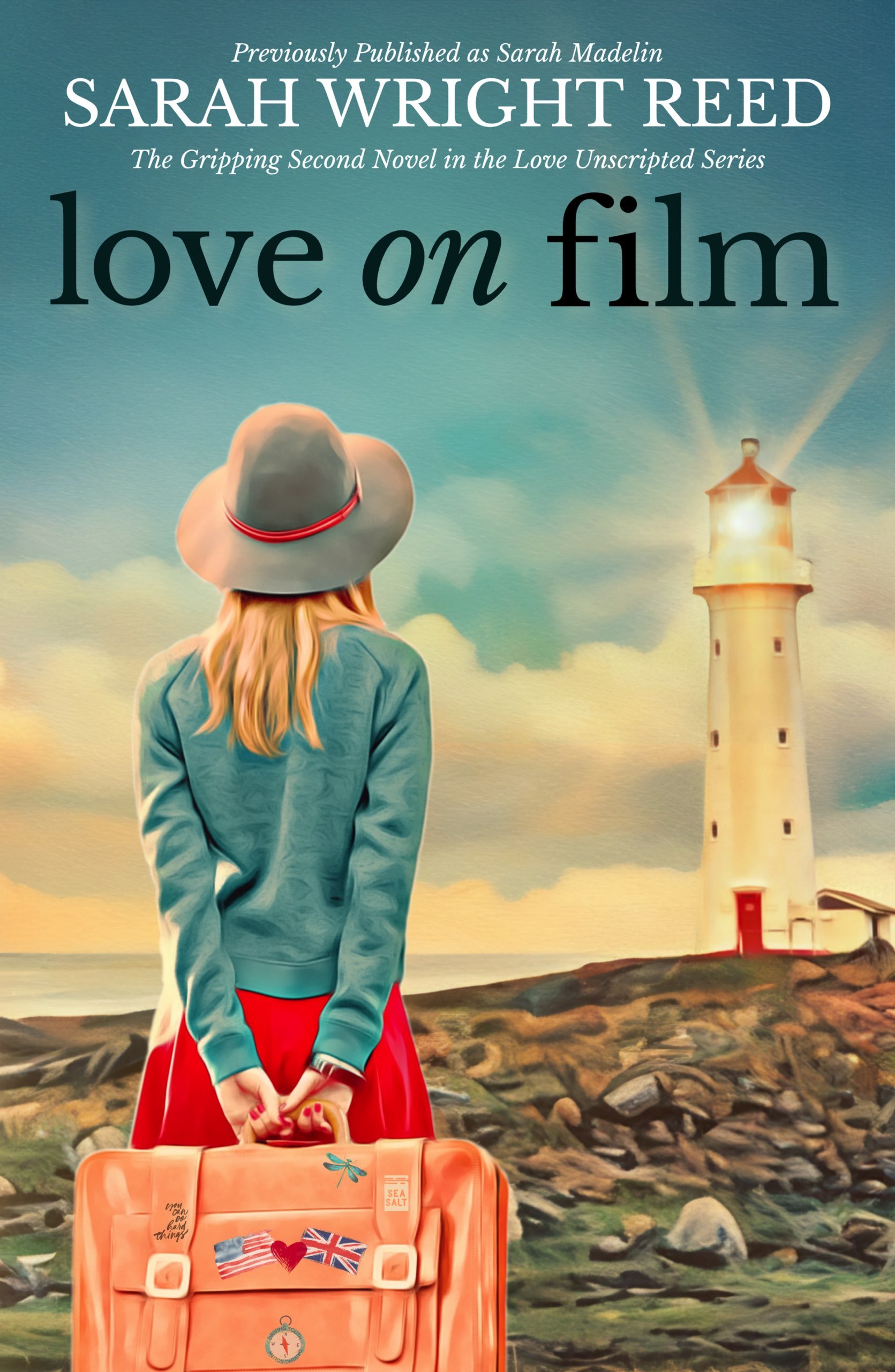 A woman with long blonde hair, wearing a wide-brimmed gray hat with a red band, blue jacket, and red skirt, stands on rocky terrain near the ocean during sunset. She holds a tan suitcase decorated with British and American flags and heart stickers. A lighthouse is visible in the background with light beams shining outward, and a cloudy sky creates a scenic seaside view.