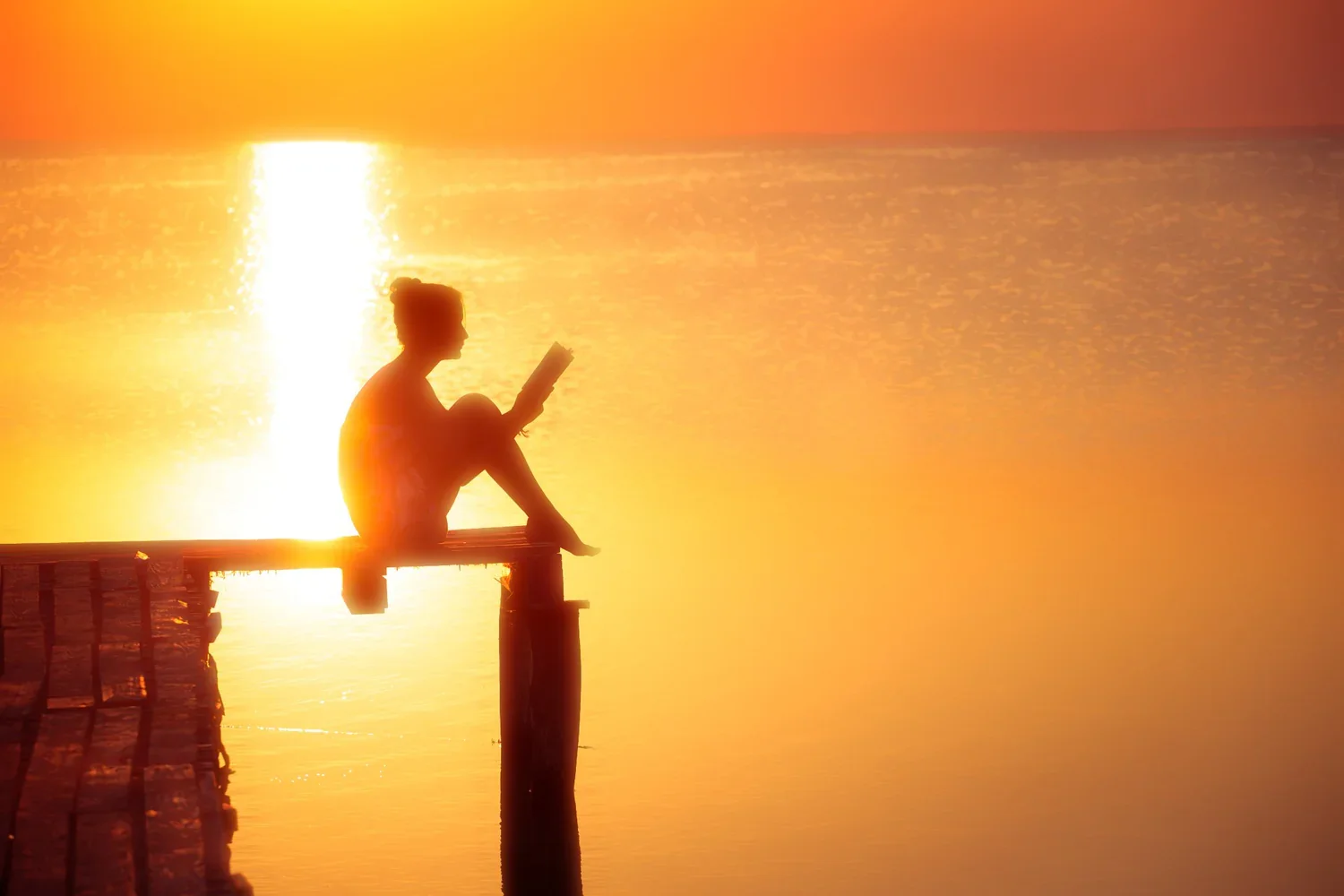 Silhouette of a woman sitting on a dock reading a book at sunset, with the sun reflecting on the water.