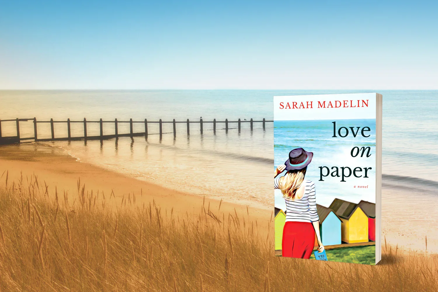 A beach scene with golden grass, a sandy shoreline, a wooden pier extending into the calm ocean, and a clear blue sky. A paperback book titled 'Love on Paper' by Sarah Madelin stands upright on the grass near the shore, featuring a woman with blonde hair, a wide-brimmed hat, and a striped shirt, holding a book and looking toward the water.