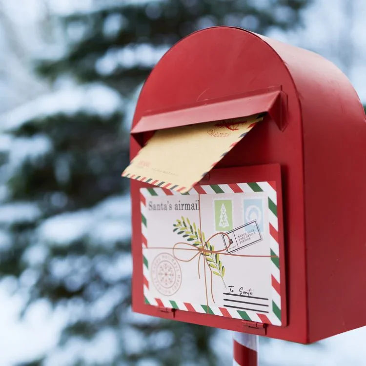 Red mailbox with a letter and Christmas card inside, set outdoors with snow and blurred trees in the background.