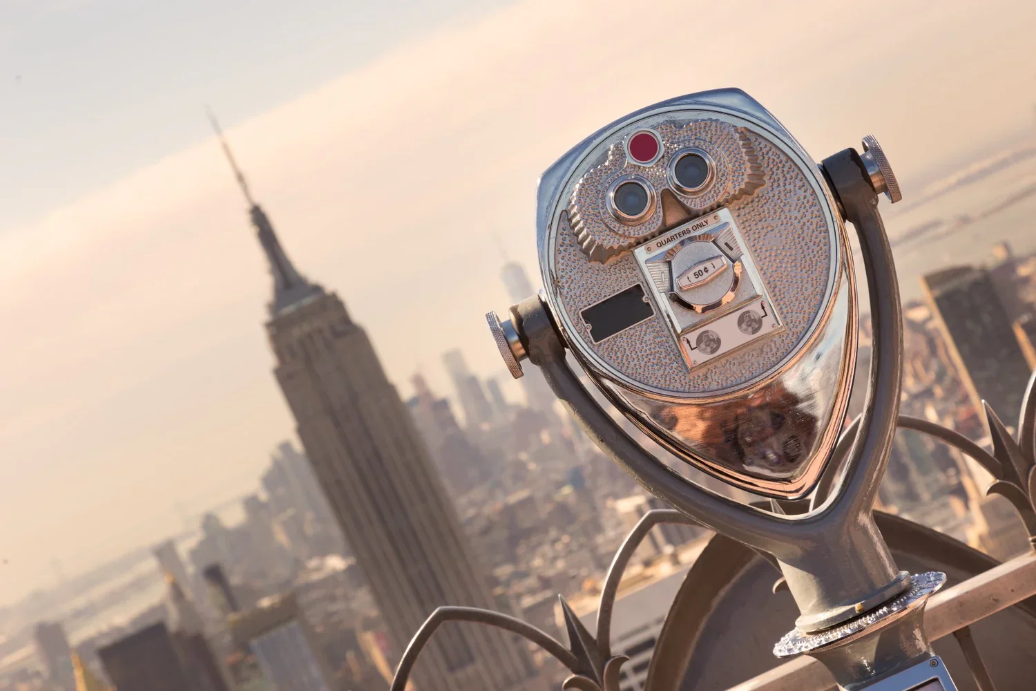Coin-operated viewing binoculars on a rooftop with a view of the Empire State Building in New York City during sunset.