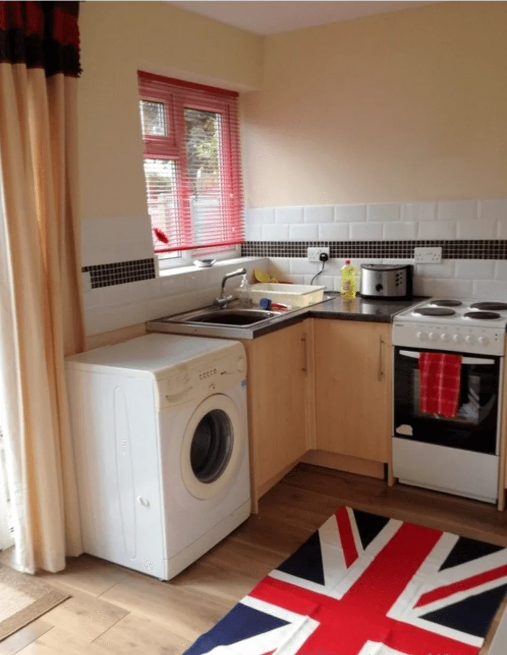 A small kitchen with a washing machine, a sink, a stove, and a toaster, decorated with UK flag-themed items and red accents.