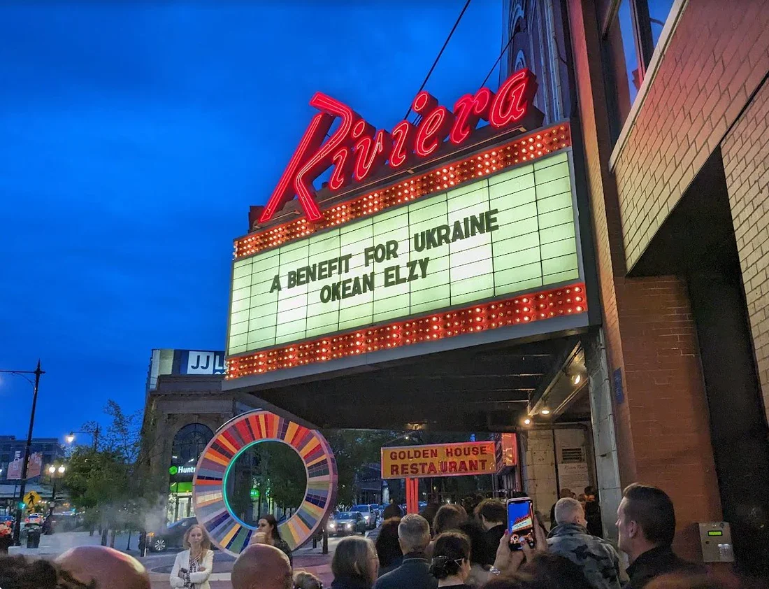 A crowd of people gathered in front of the Rivier movie theater sign which displays a message about Ukraine support. The theater marquee is lit up at dusk with a colorful circular sculpture and a sign for Golden House restaurant nearby.