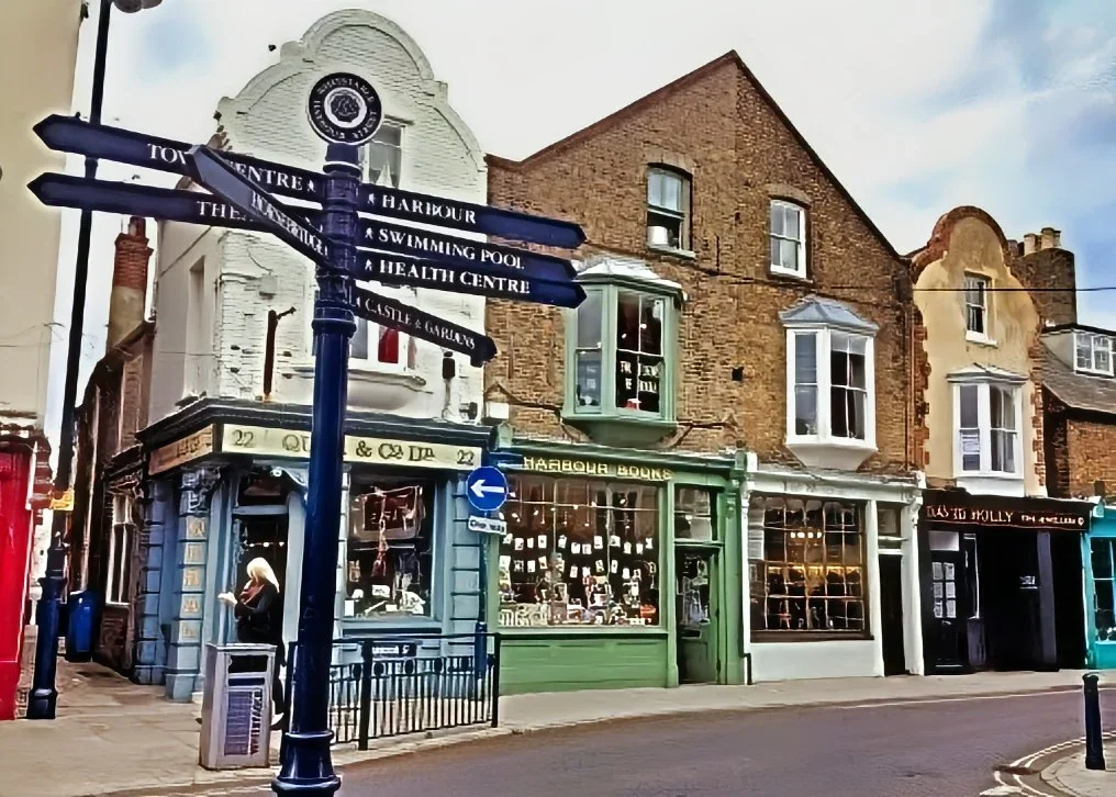 Street view of historic brick and painted buildings with various shopfronts and a street sign pointing to attractions like the town center, harbor, swimming pool, health center, and castle gardens.