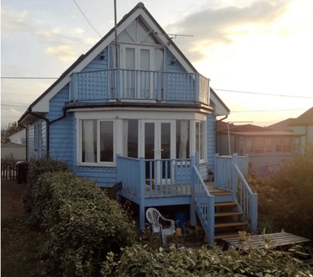 Blue house with a small balcony, front porch, and stairs, surrounded by bushes, during sunset.