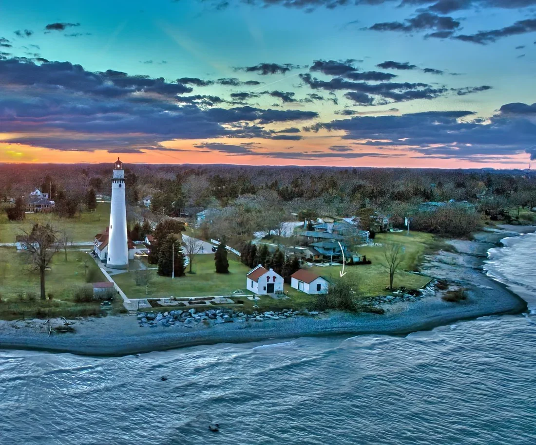 A coastal scene during sunset with a lighthouse near the shoreline, surrounded by houses and trees under a cloudy sky.