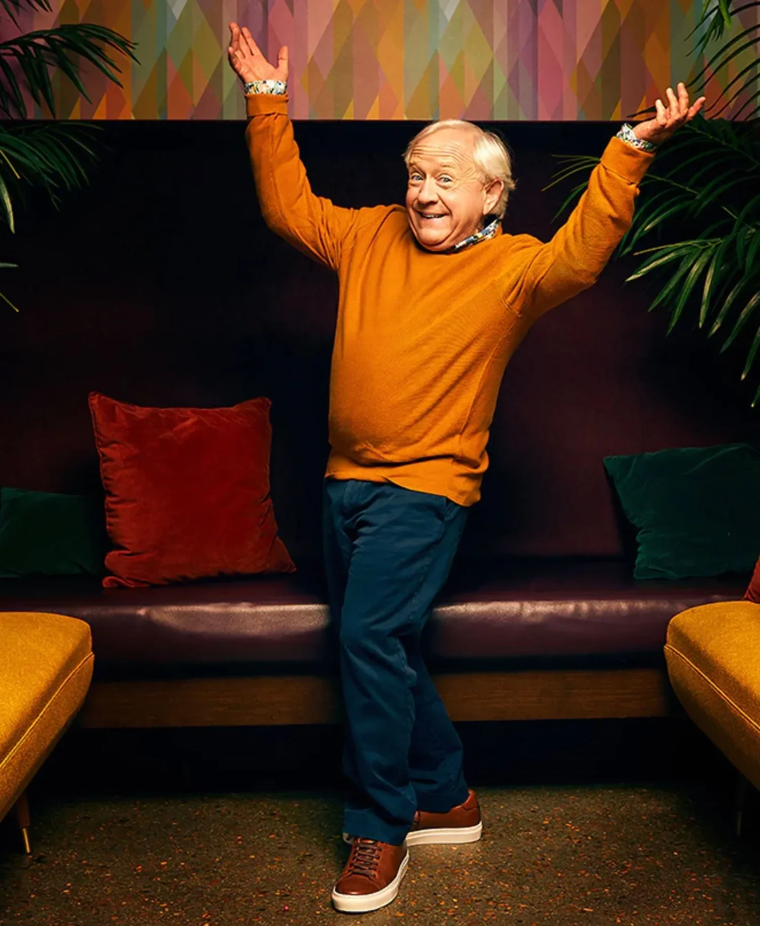 A cheerful man with white hair posing with arms raised in a colorful, modern living room with patterned wallpaper and velvet cushions.