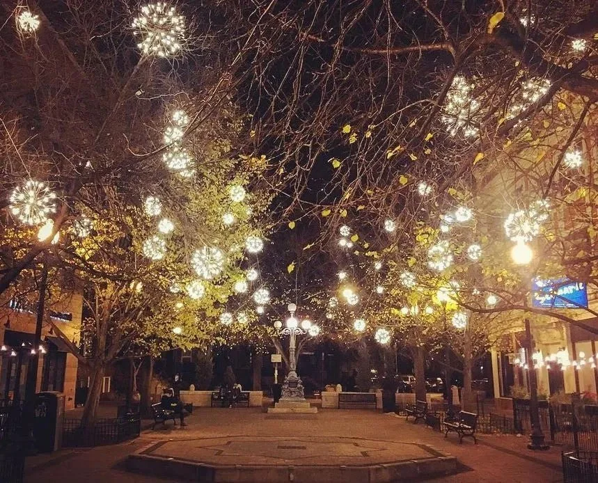 Nighttime city square decorated with hanging illuminated spherical lights, trees with sparse leaves, benches, and street lamps.