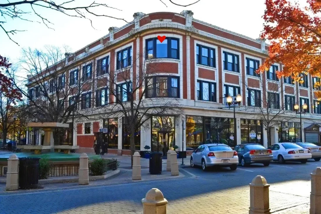 A city street scene featuring a historic red brick building with multiple windows, trees with autumn foliage, parked cars, and a small green park with a fountain.