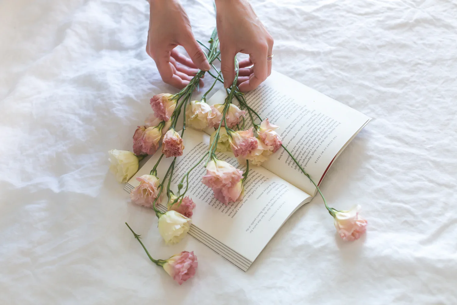 Hand arranging pink and white carnations on an open book placed on a white textured fabric surface.