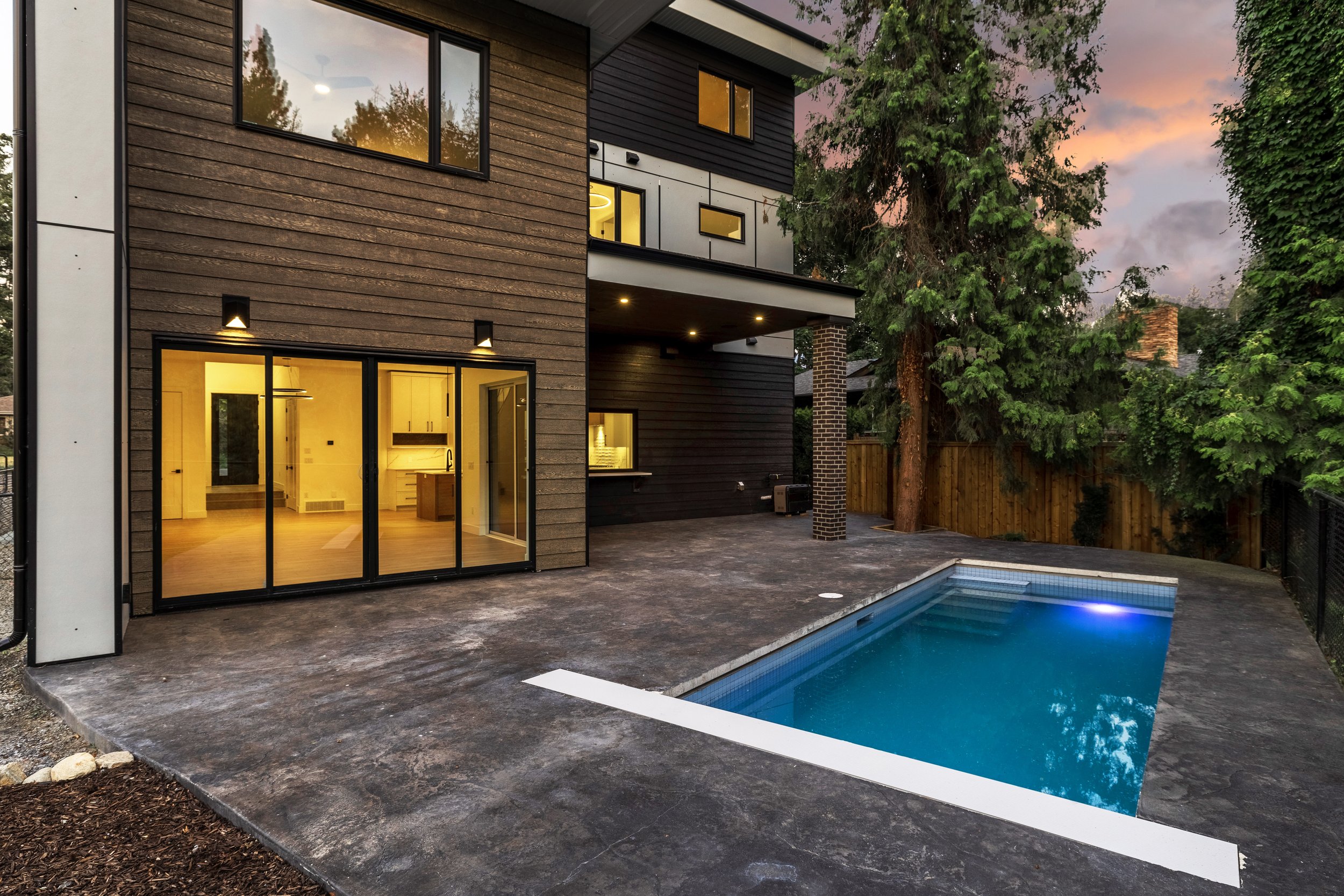 A modern house with large glass sliding doors overlooking a backyard with a swimming pool, trees, and a wooden fence at sunset.