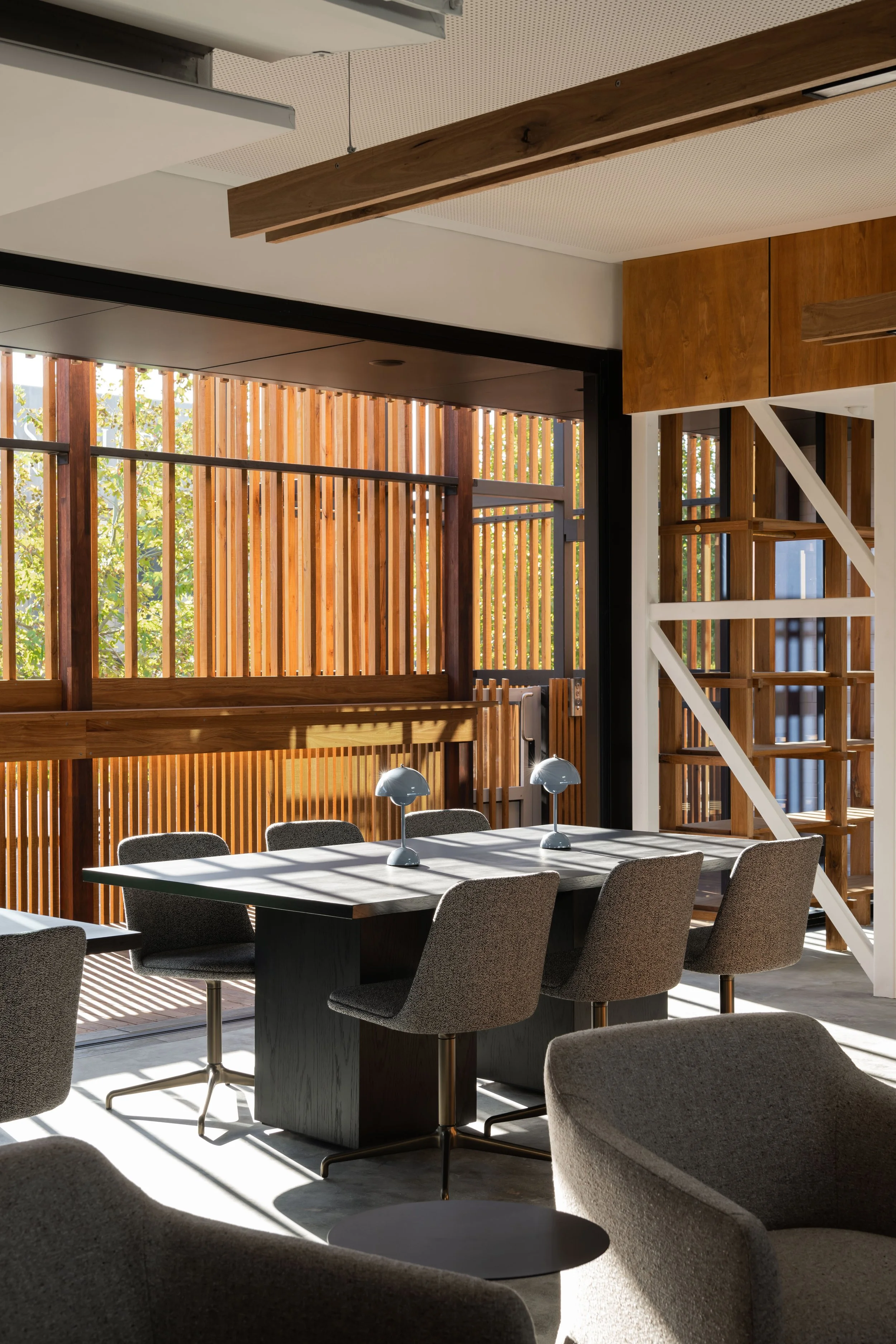 A modern office or conference room with a black table, gray chairs, and small lamps on the table, with wooden slat walls and natural sunlight streaming in.