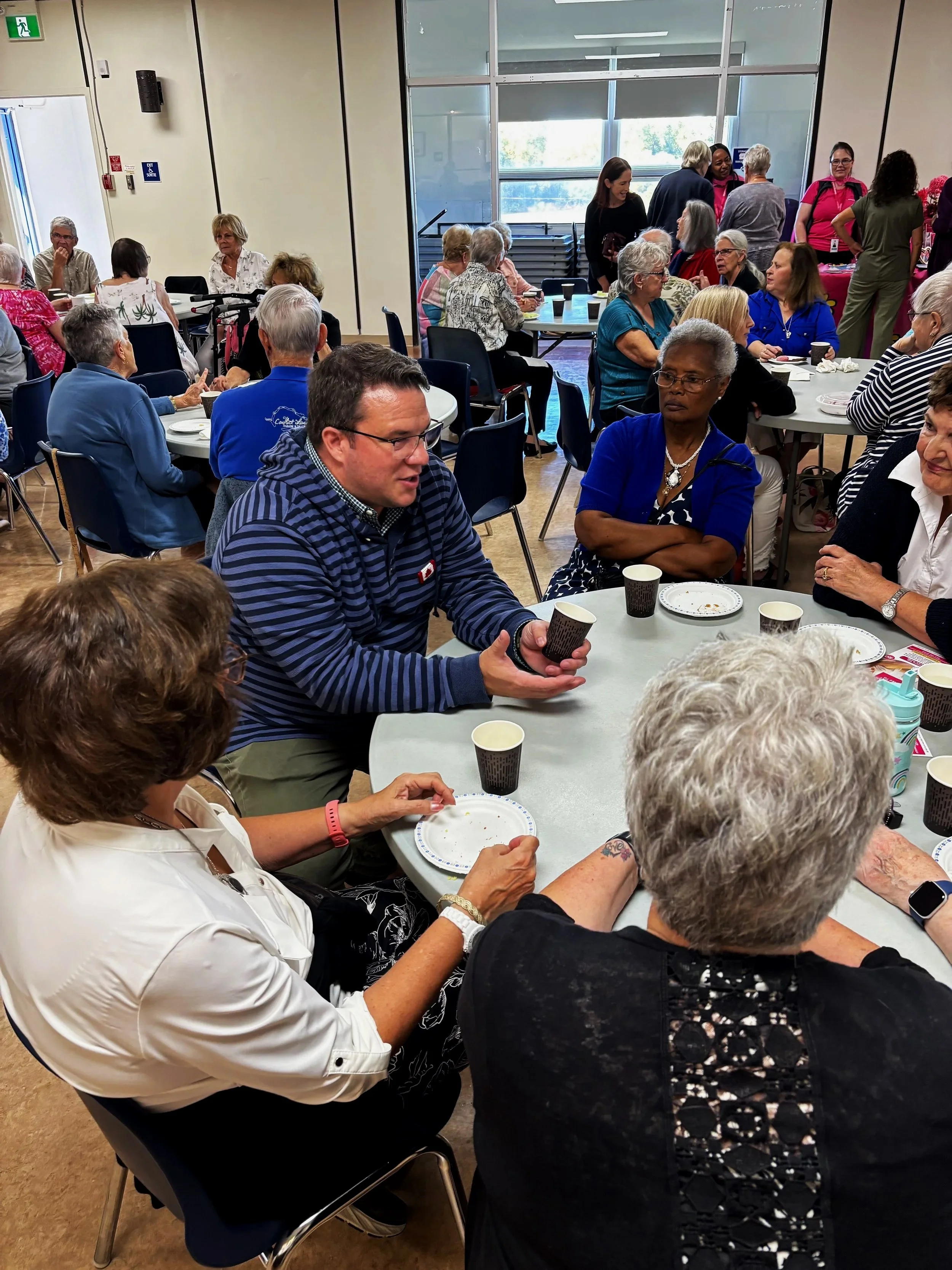 Group of senior women and some men gathering around tables in a community room, engaging in conversation with Matt Luloff. Some women are sitting, others standing, with plates, cups, and utensils on the tables.