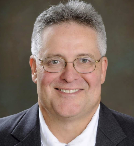 A smiling man with short gray hair and glasses, wearing a dark suit jacket over a white shirt, posing for a portrait against a neutral background.