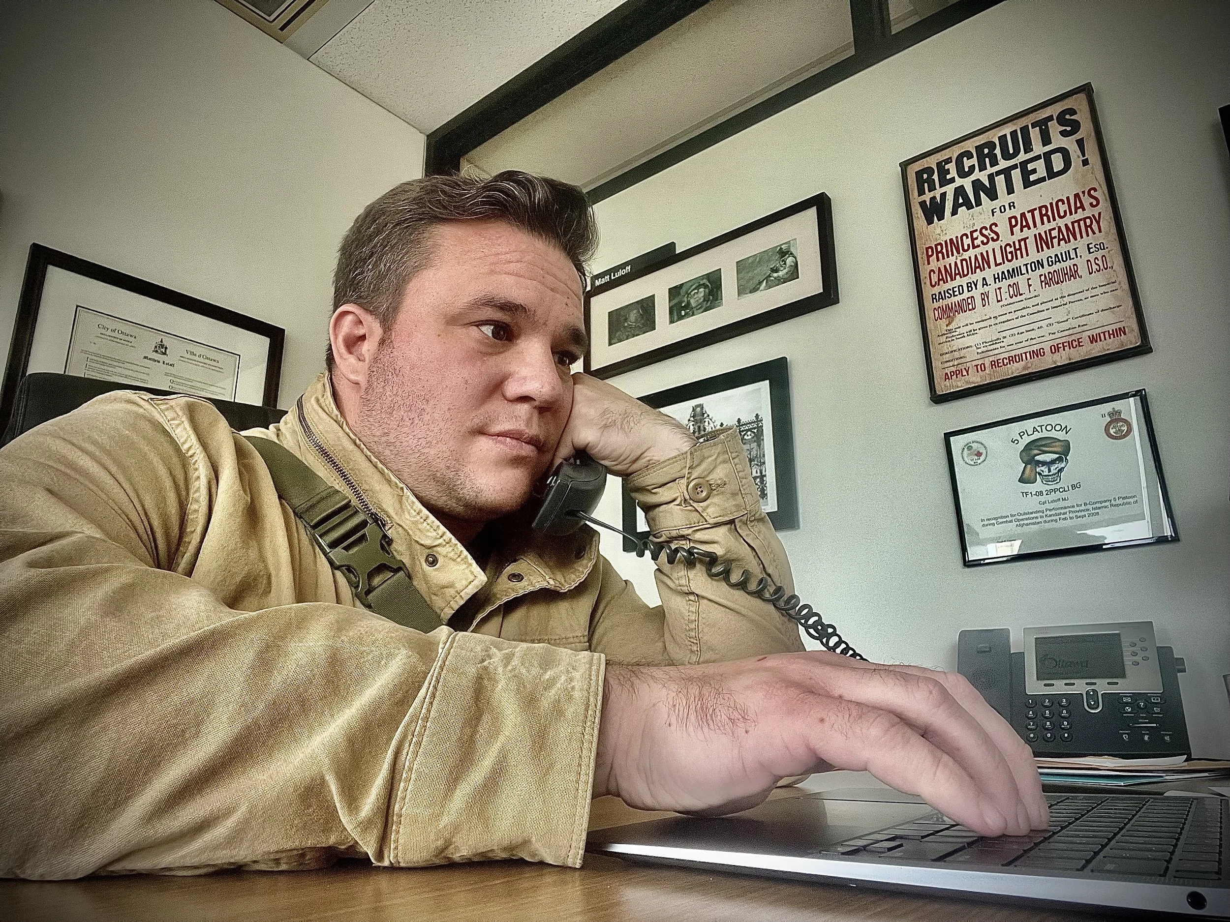 Matt Luloff talking on a landline phone while working on a laptop desk with framed documents and posters on the wall behind him.
