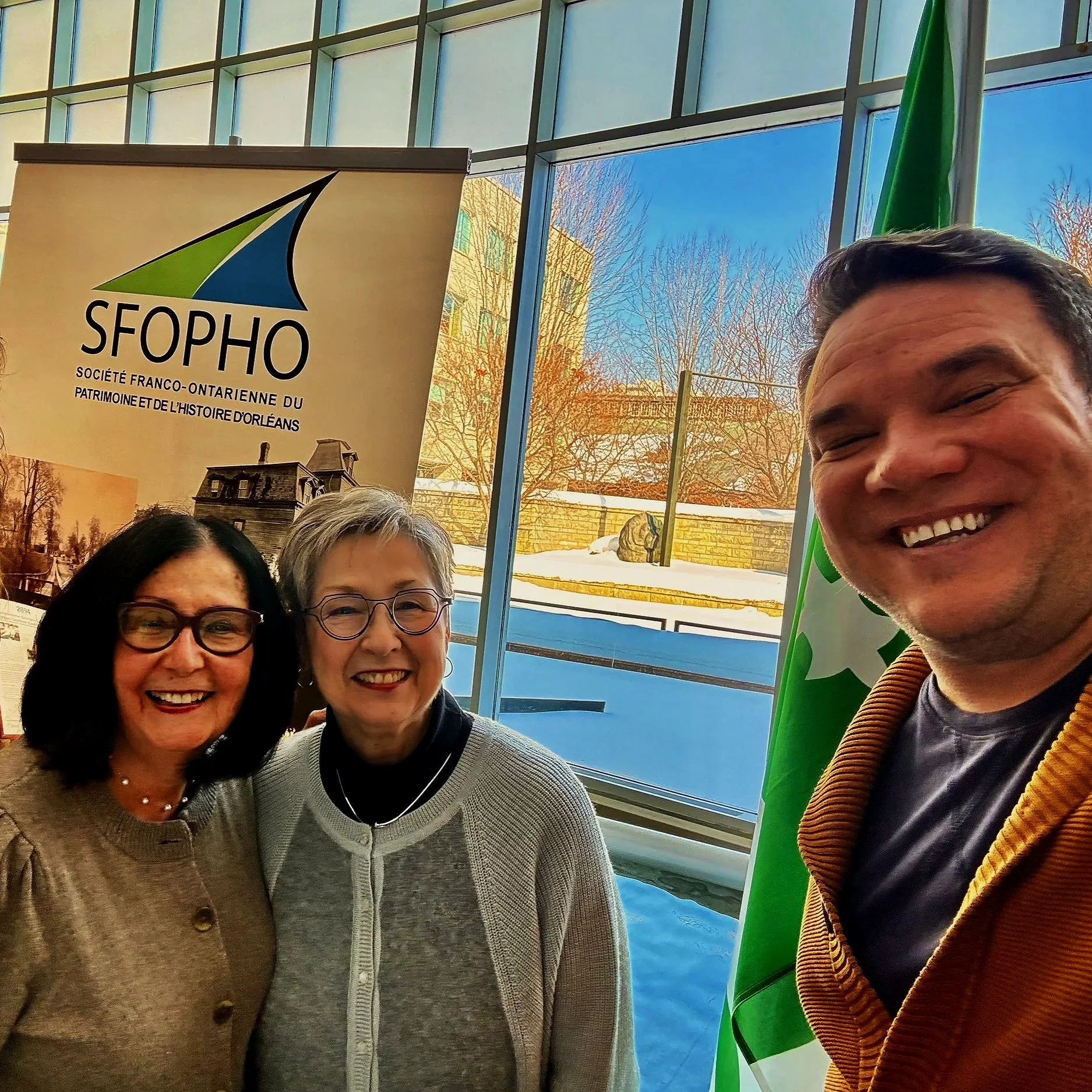 A group of smiling people are taking a selfie indoors. Behind them, there is a large window or glass wall showing trees and a building outside. A banner with a logo and text in French is also visible.