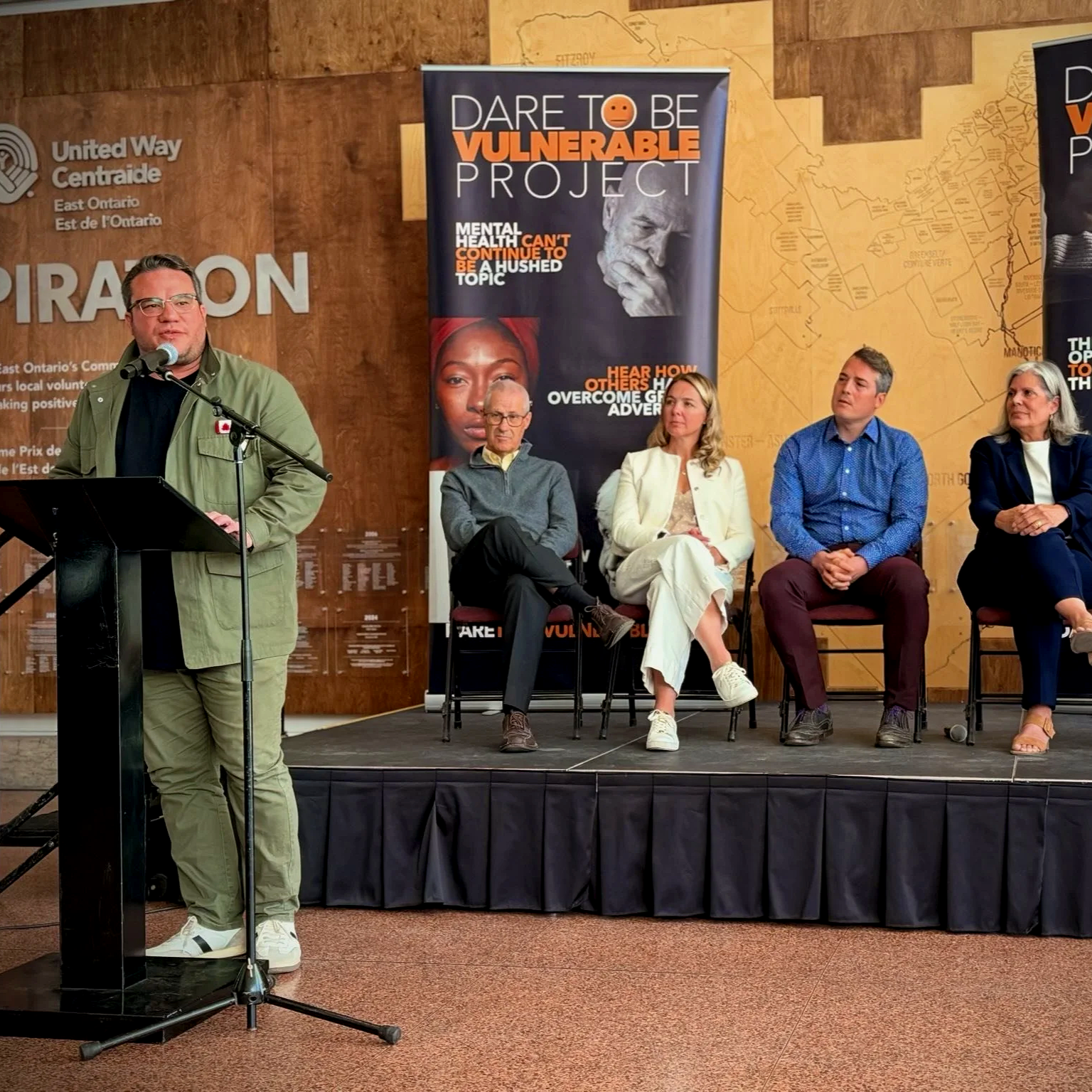 Matt Luloff is speaking at a podium with a microphone at a panel discussion. Four people are seated behind him on a stage, with a large banner about mental health and vulnerability placed behind them. The background features wood paneling and a map.