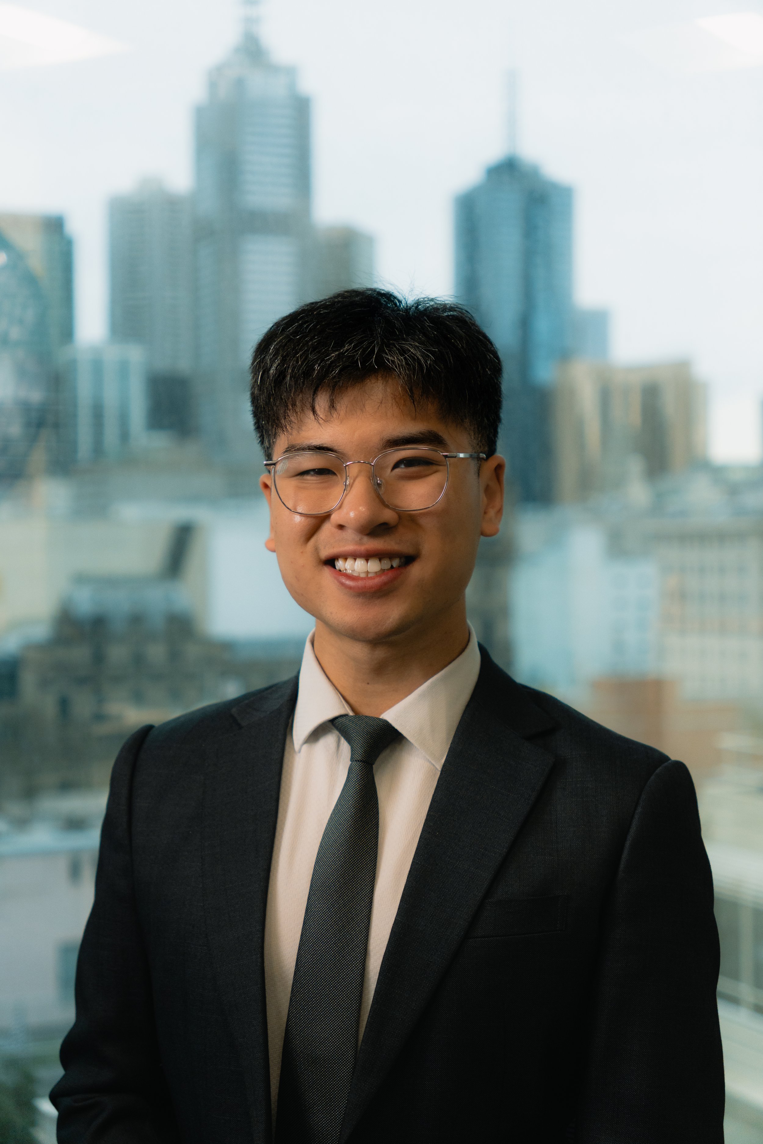 Young man with glasses wearing a suit and tie, smiling in front of a cityscape with tall buildings.