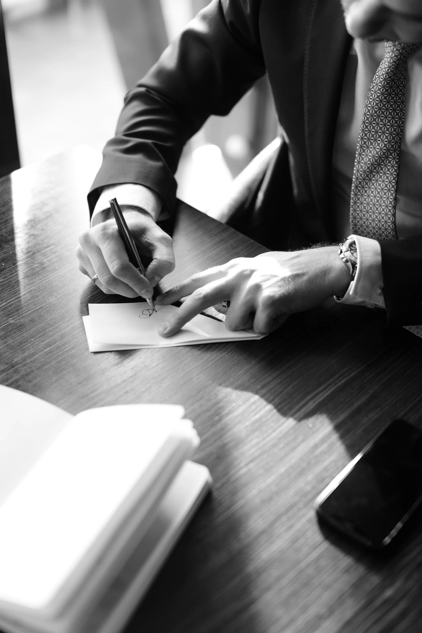 A man in a suit signing a document on a wooden table with a pen.