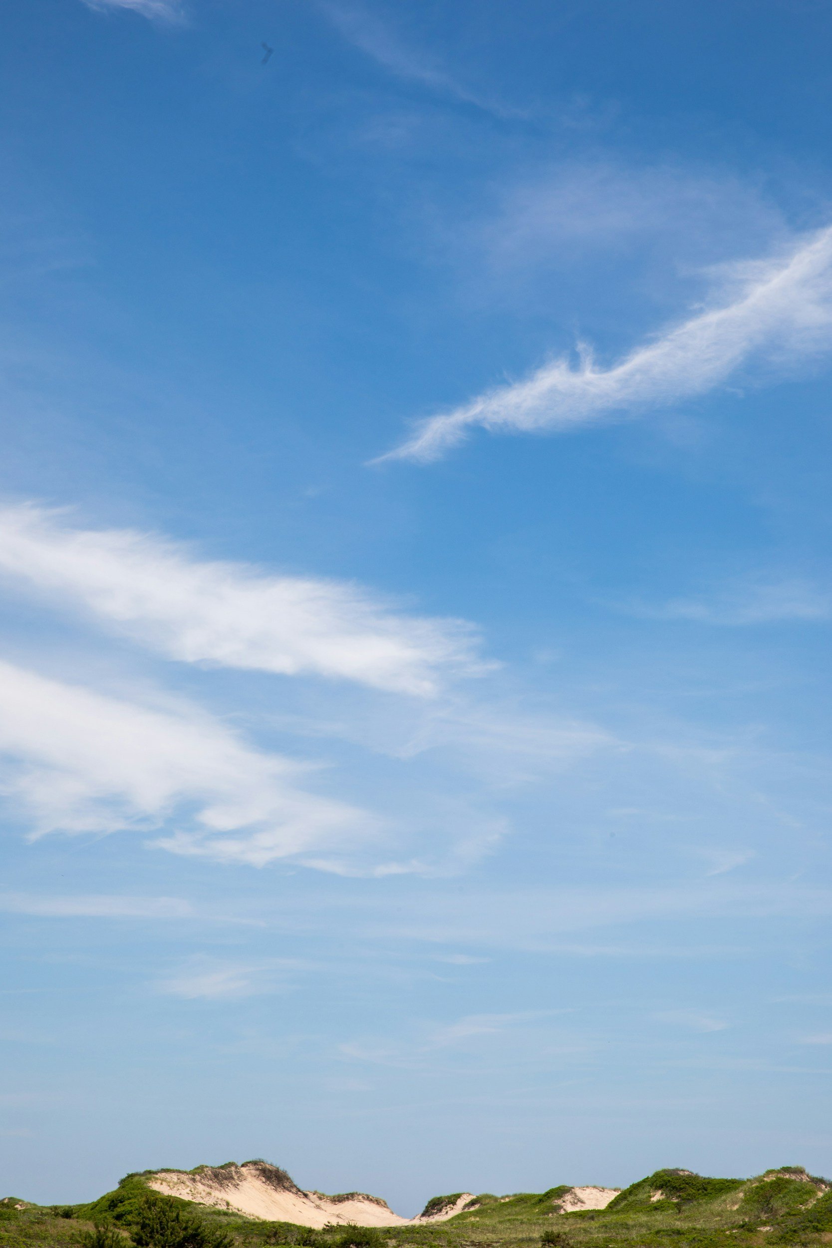 A landscape with sandy dunes at the bottom, covered in green vegetation, under a bright blue sky with scattered wispy white clouds.
