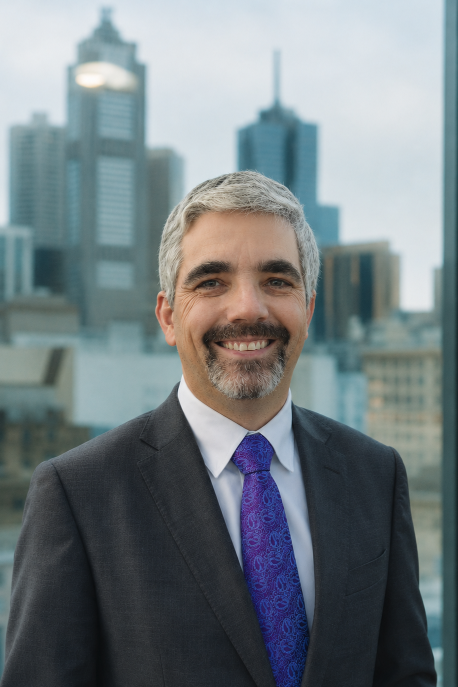 Smiling middle-aged man with gray hair and beard, wearing a dark suit, white shirt, and patterned blue tie, standing in front of a cityscape with tall skyscrapers.