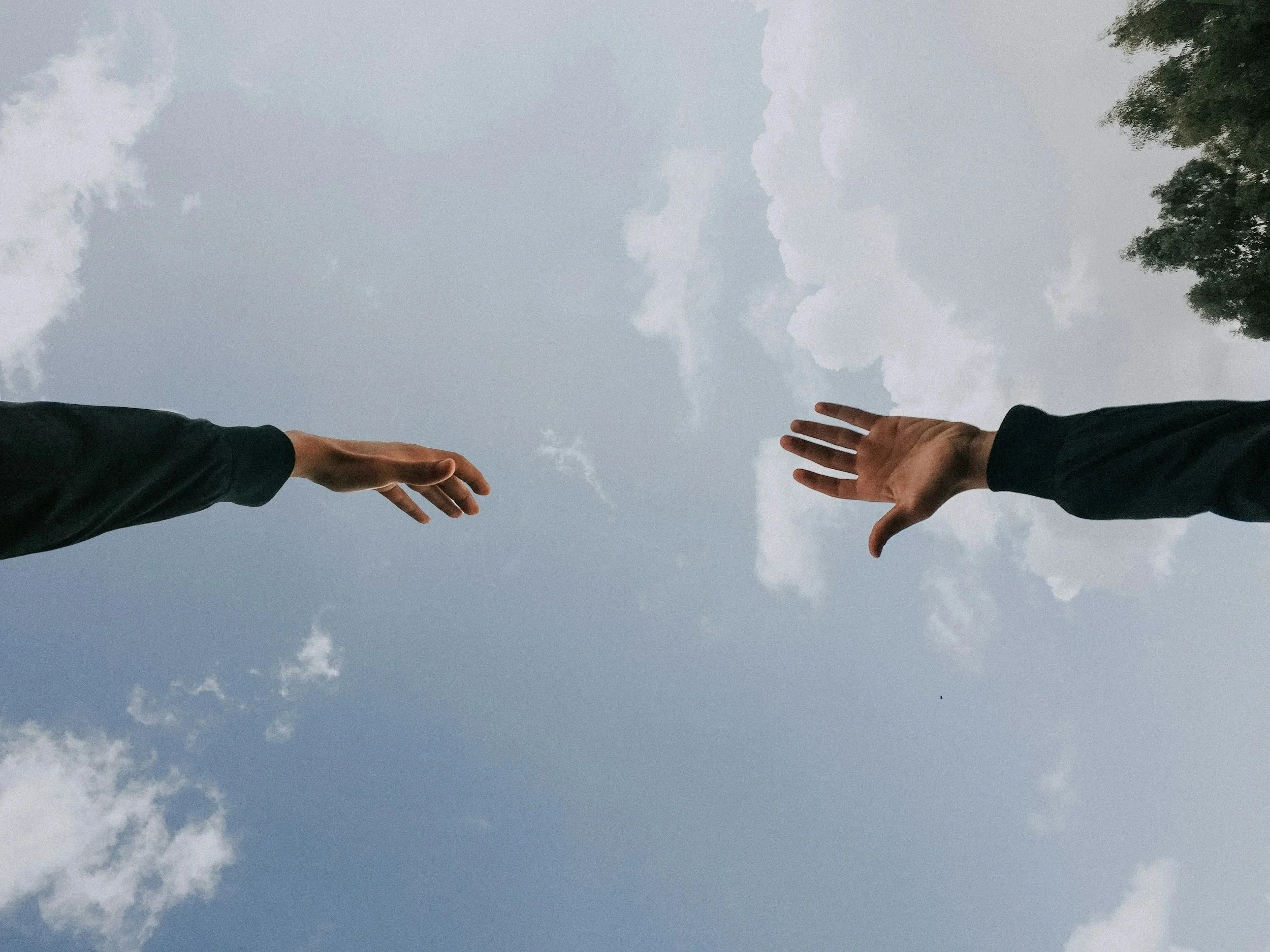 Two people reaching their hands toward each other in the sky, with clouds and trees in the background.