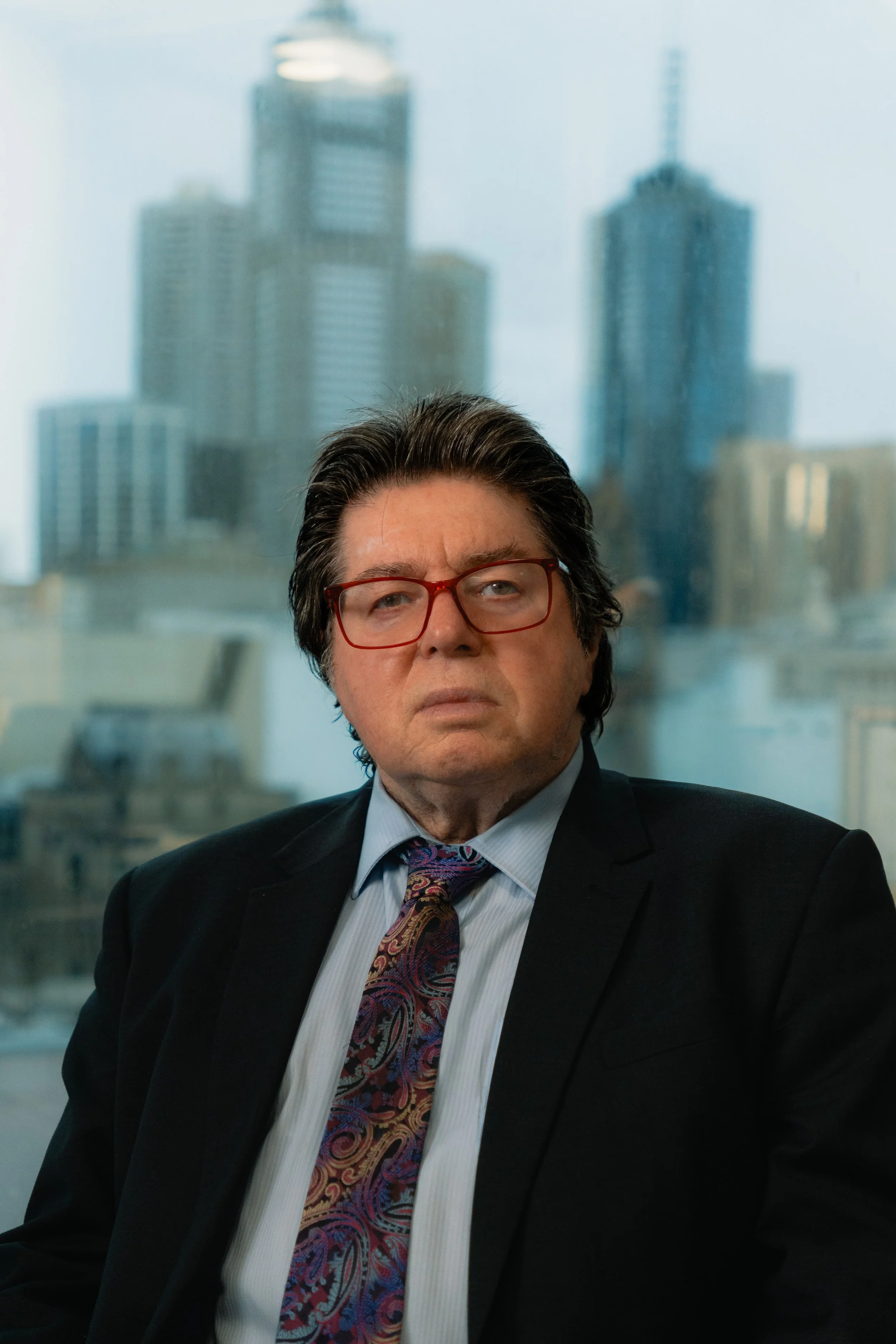 A middle-aged man with dark hair and glasses, wearing a black suit, light blue shirt, and a colorful paisley tie, sitting in an office with a city skyline view.
