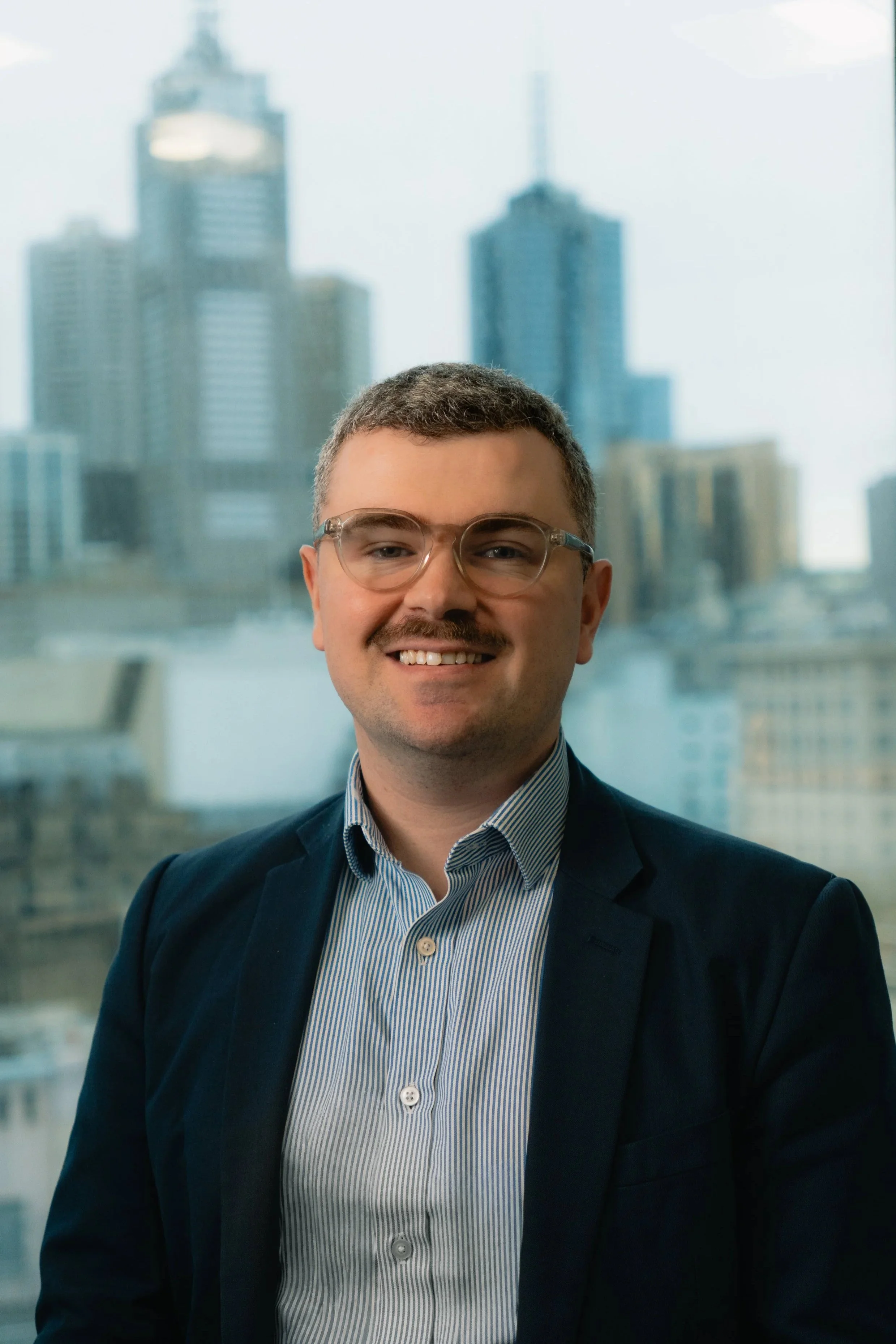 A man wearing glasses and a dark suit jacket, smiling, standing in front of a large window with a city skyline of tall buildings in the background.