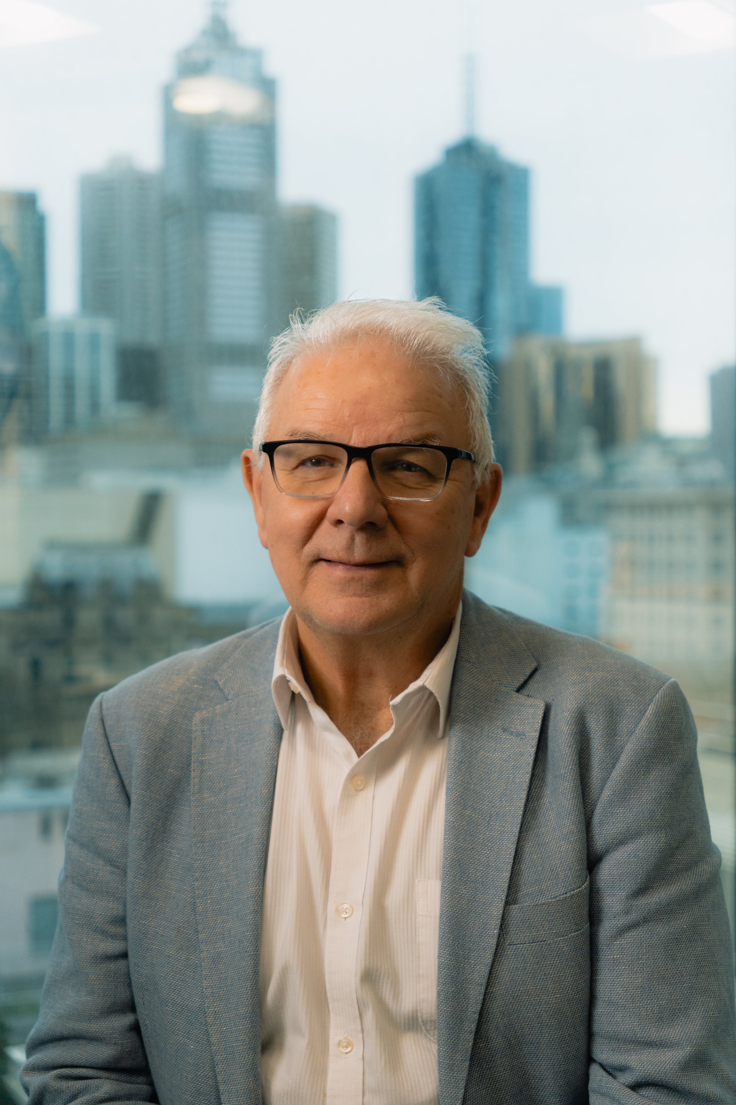 A middle-aged man with gray hair and glasses wearing a gray suit jacket and white shirt, smiling, with a cityscape of tall buildings in the background.
