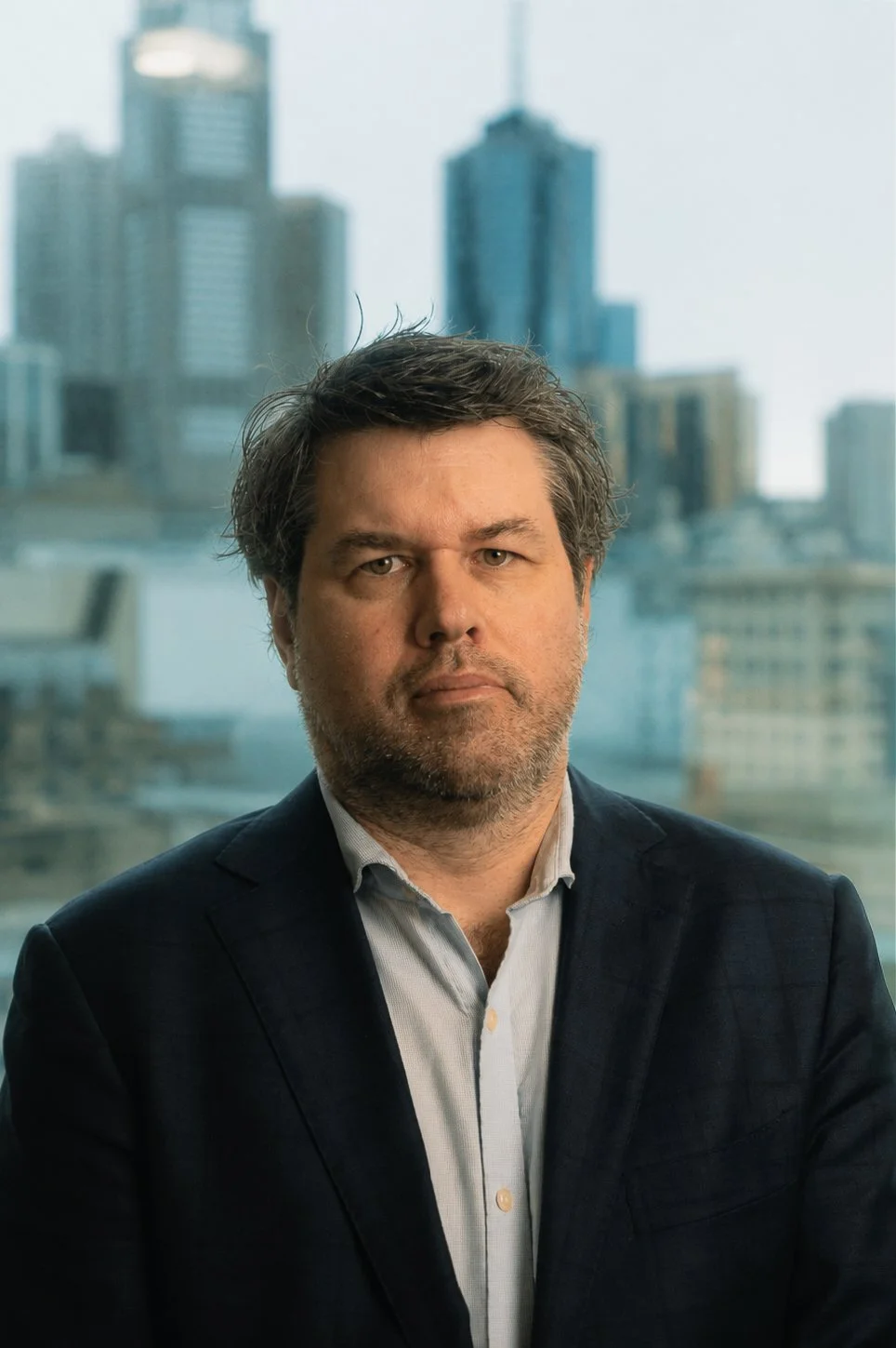 A man with dark hair and a beard in a suit in front of a city skyline with tall buildings.