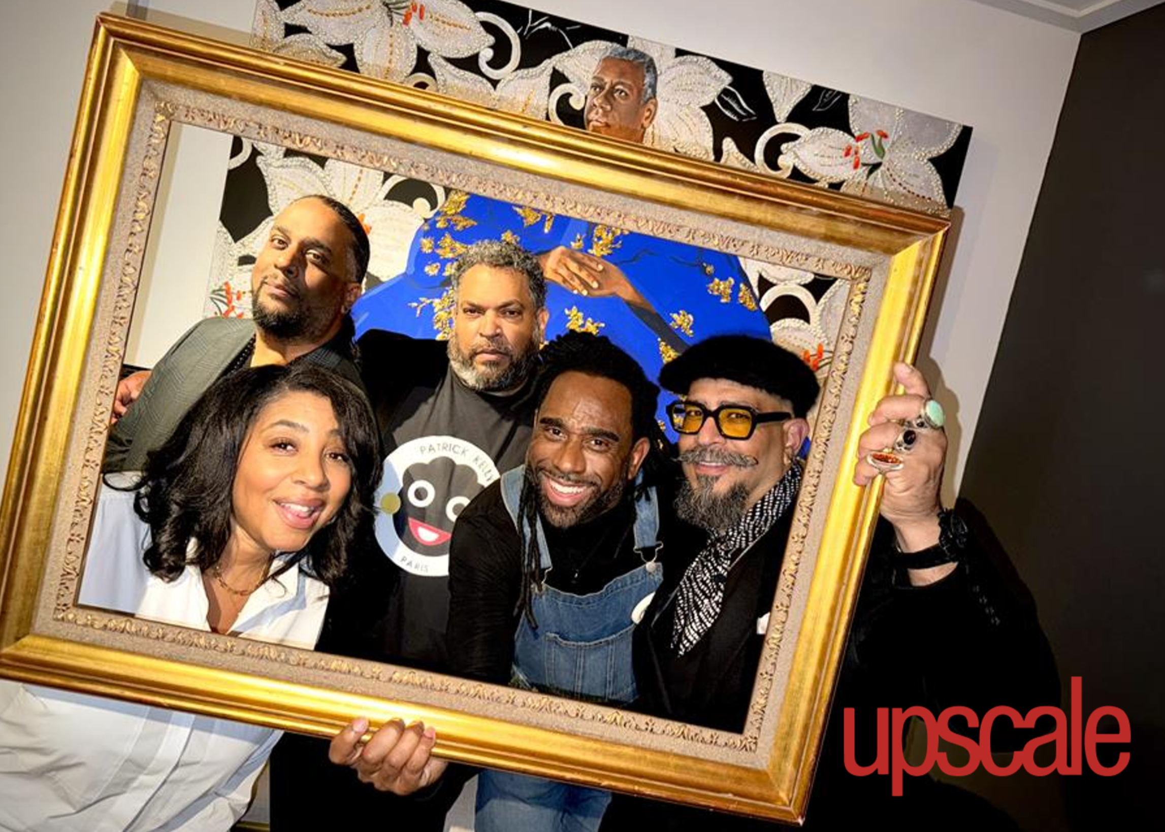 A group of people posing behind a large framed artwork. The artwork features a portrait of a man with a dark background decorated with white and gold floral patterns, and a woman in a blue outfit with gold accents is part of the artwork. The individuals are smiling and appear to be at an art exhibition or gallery.