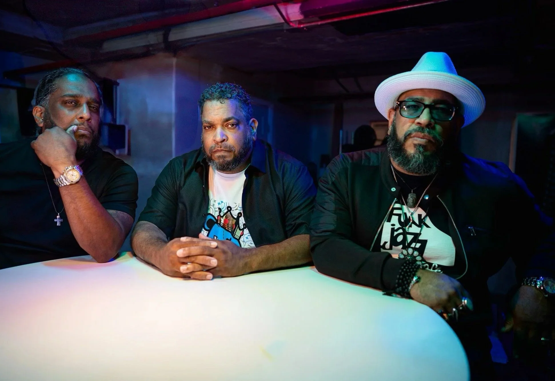 Three men sitting at a table in a dimly lit room with colored lighting, facing the camera. The man on the left has a beard and moustache, wearing a black t-shirt and a silver watch, with his hand on his chin. The man in the middle has a beard, short curly hair, wearing a black shirt over a graphic t-shirt. The man on the right has a beard, glasses, wearing a white hat, black glasses, and a black jacket with a white graphic t-shirt underneath.