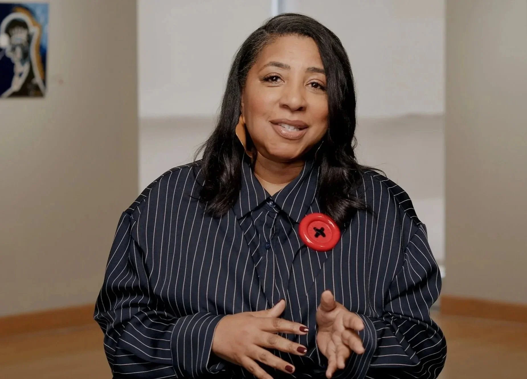 A woman with dark hair wearing a black and white pinstripe shirt with a red button near her collar, speaking indoors with a neutral background.