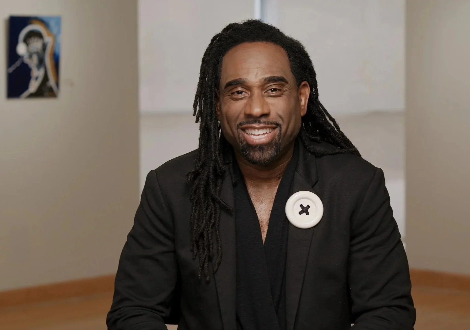 A man with long dreadlocks, wearing a black blazer with a large white button pin, smiling and sitting indoors.