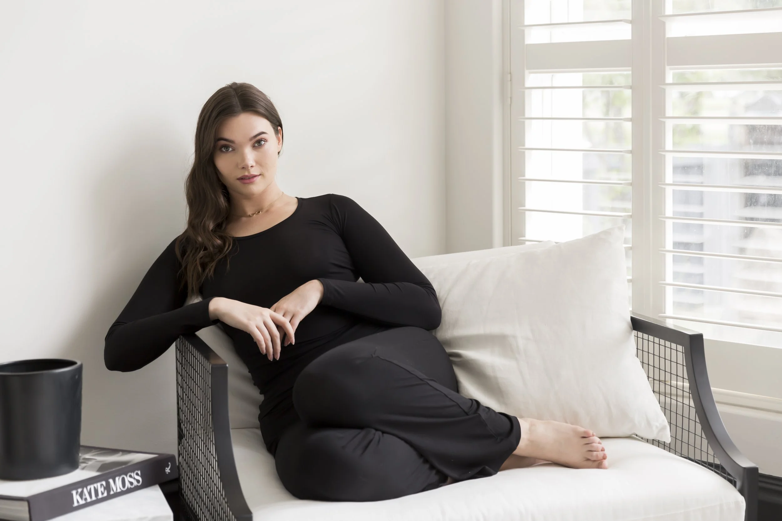 A woman with long brown hair, wearing a black top and black pants, sitting on a white outdoor lounge chair near a window with white shutters, in a bright room.