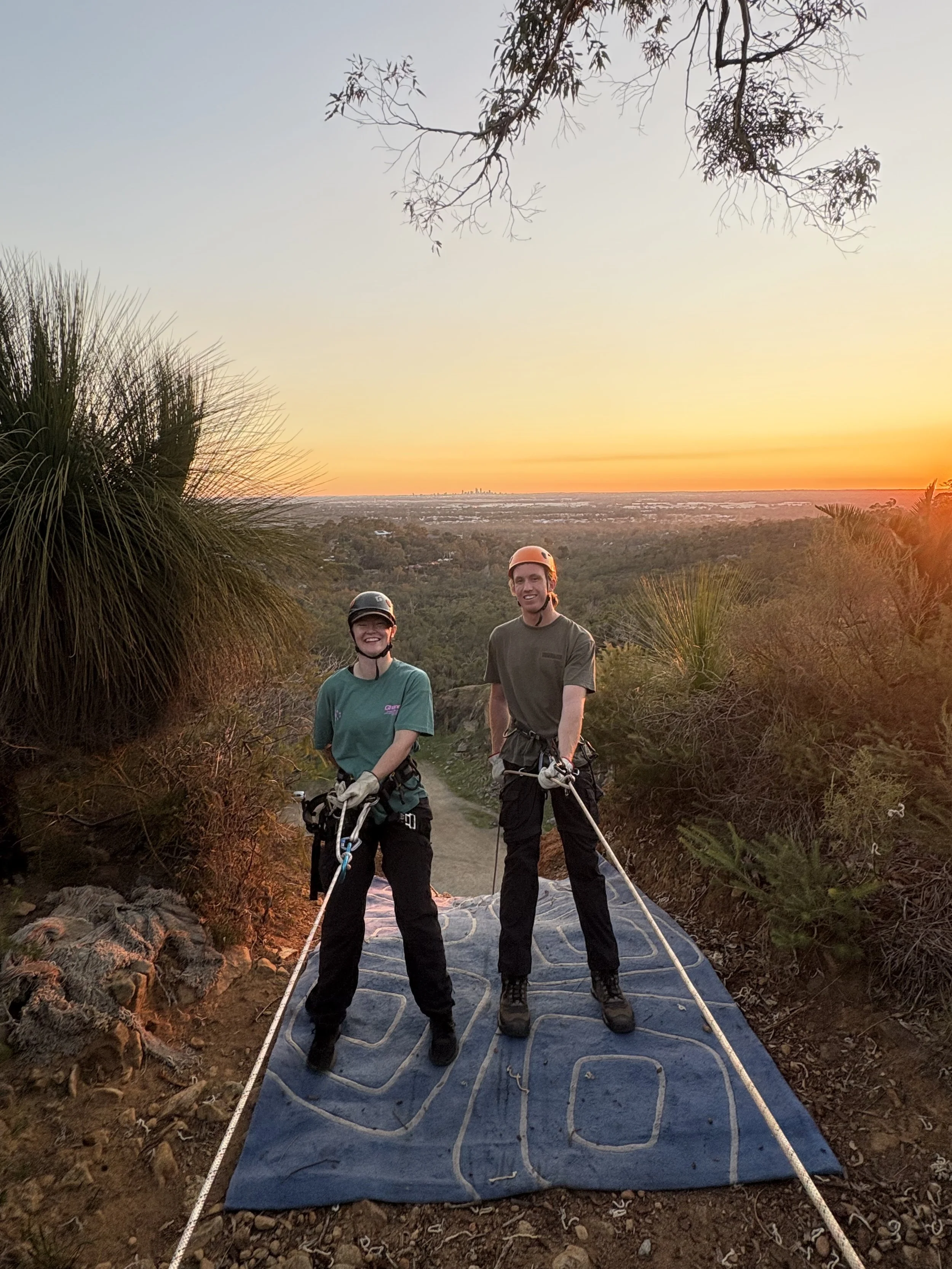 Two people wearing helmets and harnesses standing on a crash pad outdoors during sunset, surrounded by trees and vegetation.