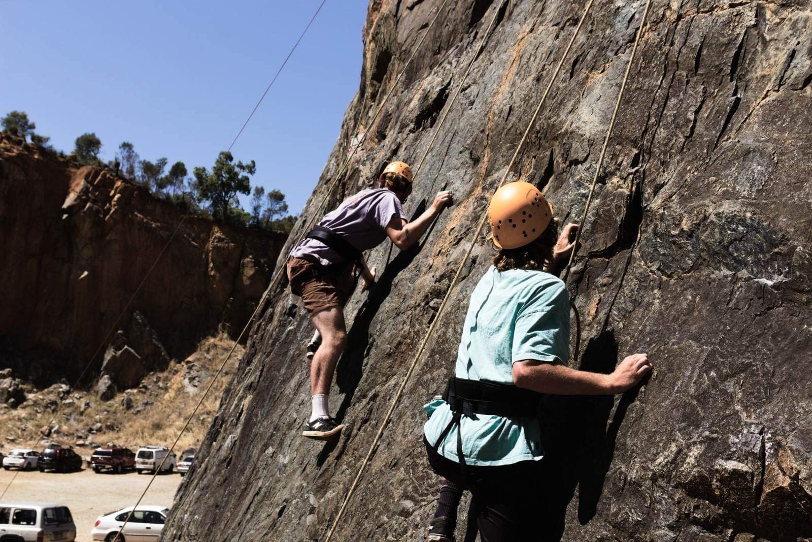 Two people rock climbing on a vertical rock face, wearing helmets and harnesses, with a parking lot and mountains in the background.