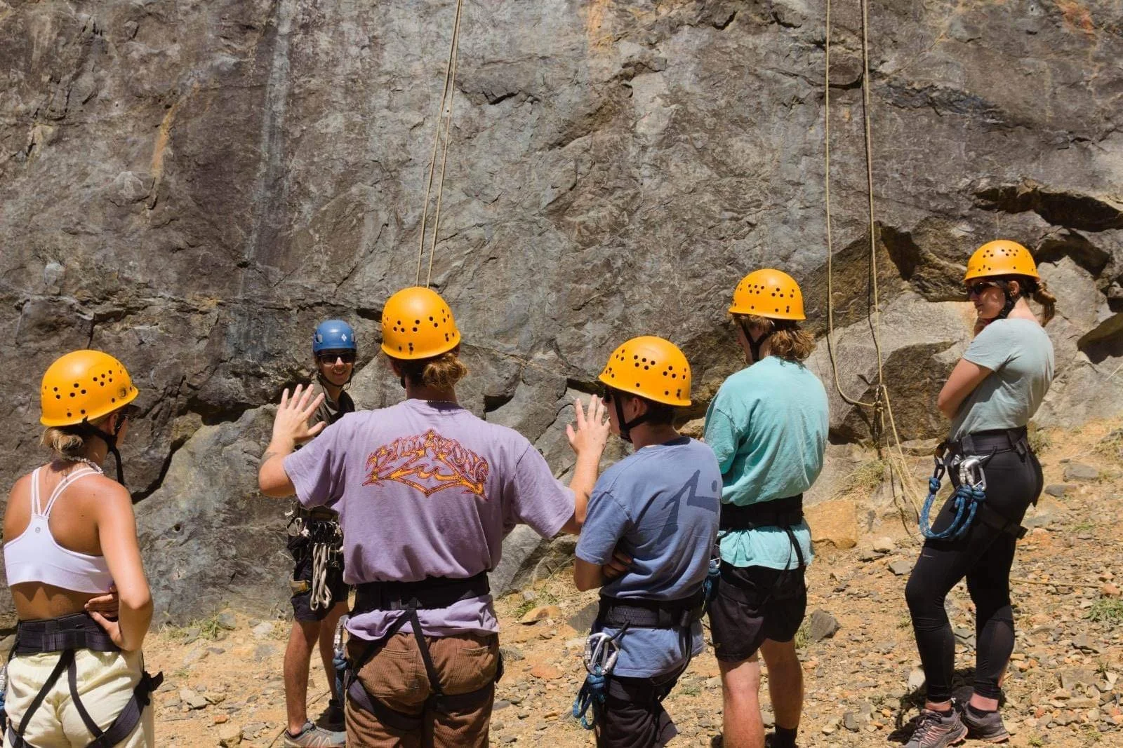 Group of people wearing safety helmets gathered at the base of a rocky cliff, preparing for a rock climbing or rappelling activity.