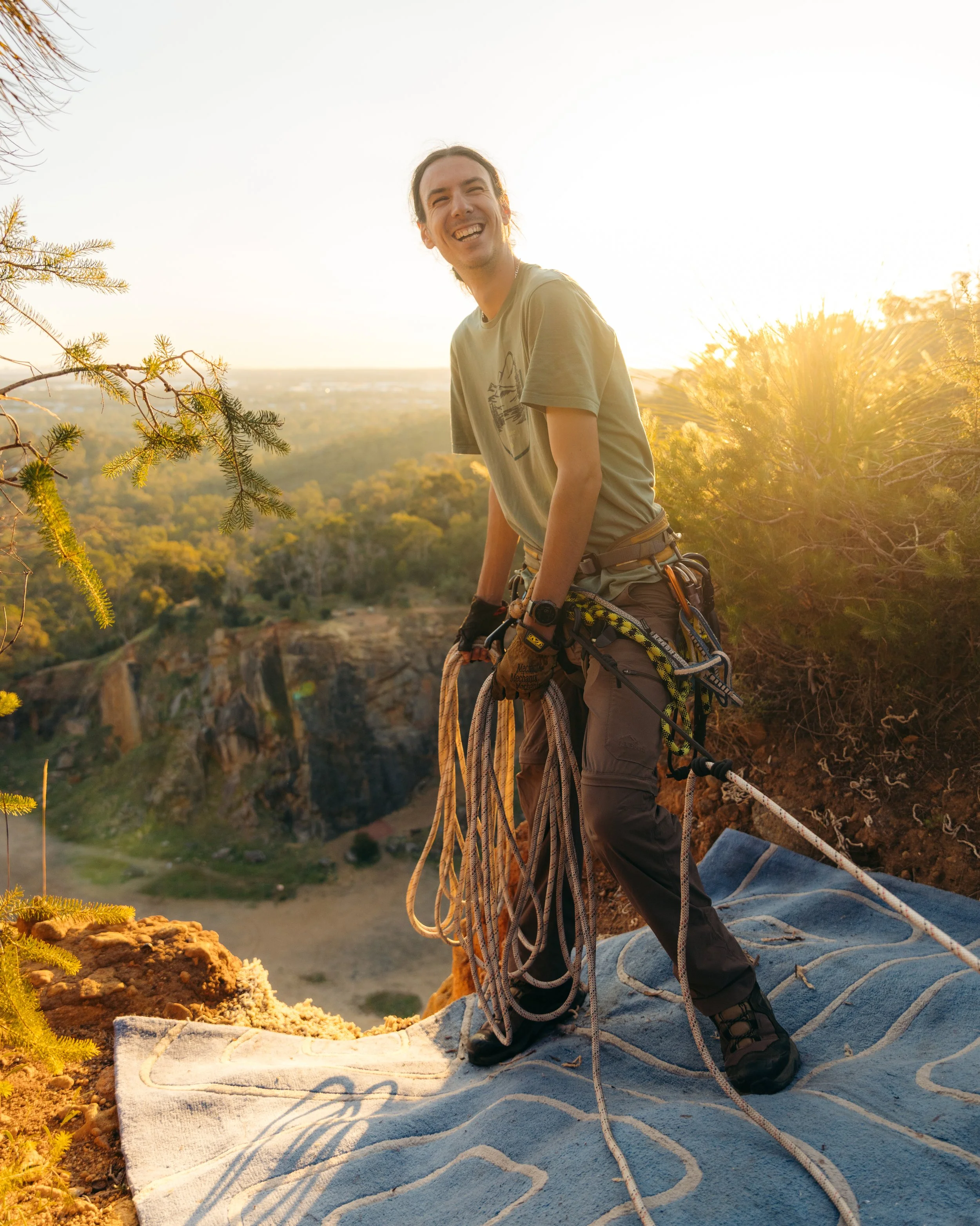 Person in outdoor climbing gear sitting on a blanket on rocky terrain, smiling at sunset with trees and landscape in background.