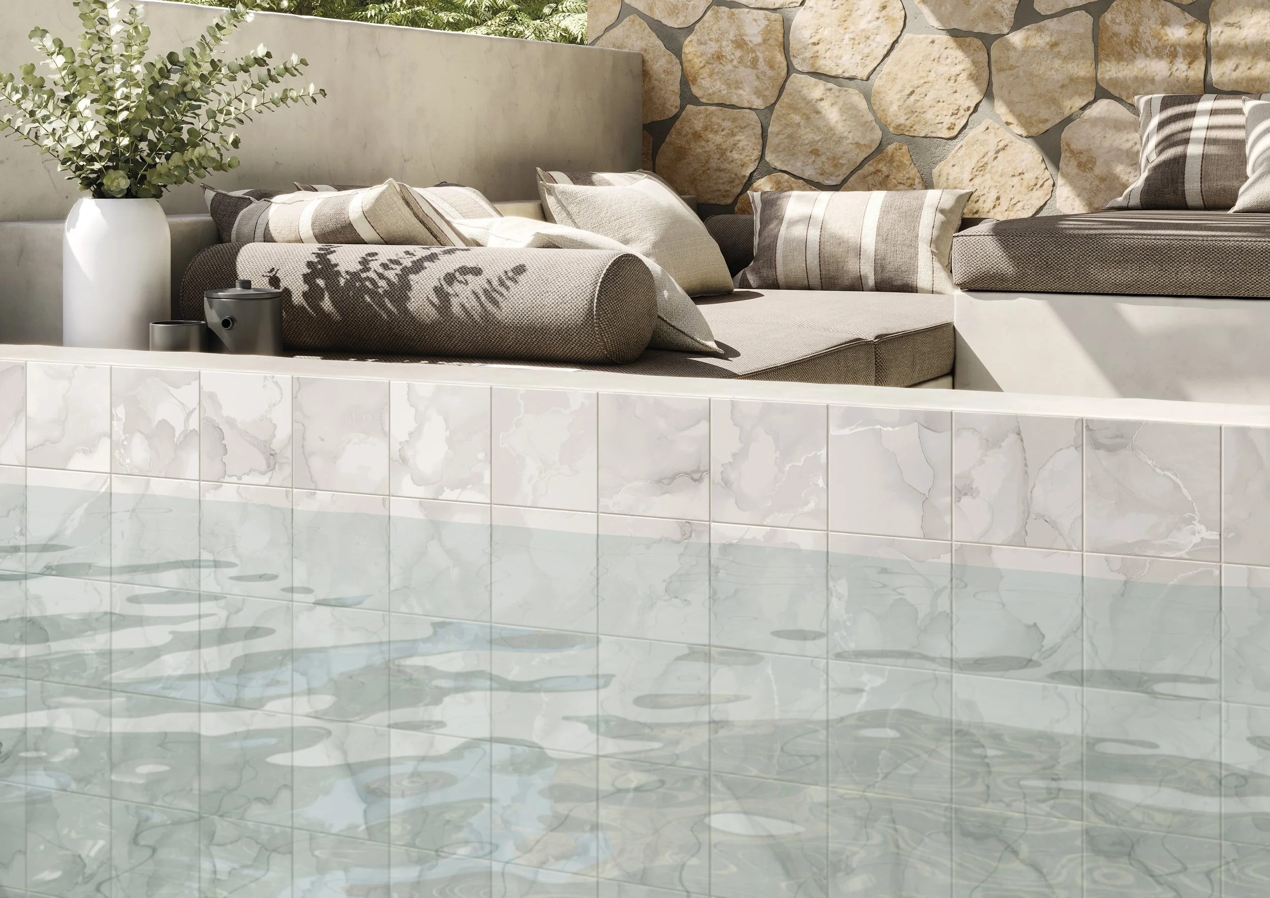 Une piscine avec de l'eau claire et de la céramique blanche, un mur en pierre naturelle en arrière-plan, et un espace détente avec un canapé et des coussins beiges et à rayures, ainsi qu'une grande vase avec des feuilles vertes.