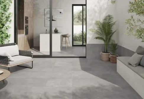 Salon moderne avec canapé blanc, plantes vertes, mur blanc, sol en béton, et porte-fenêtre donnant sur un espace extérieur