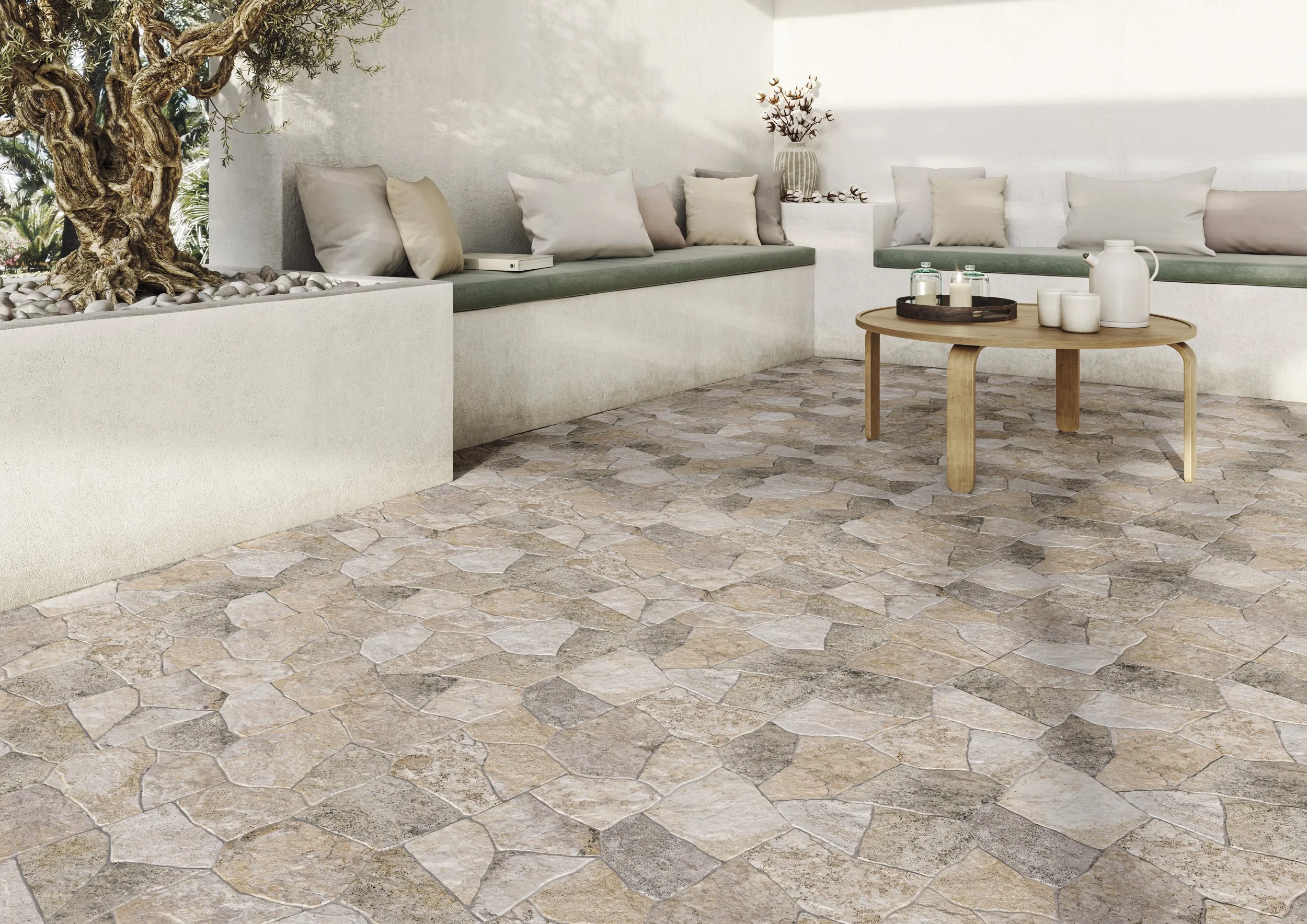Un coin salon avec un mur blanc, un canapé avec plusieurs coussins dans des tons neutres, une table en bois avec des objets décoratifs, un grand pot avec un arbre décoratif, et un plancher en pierre.