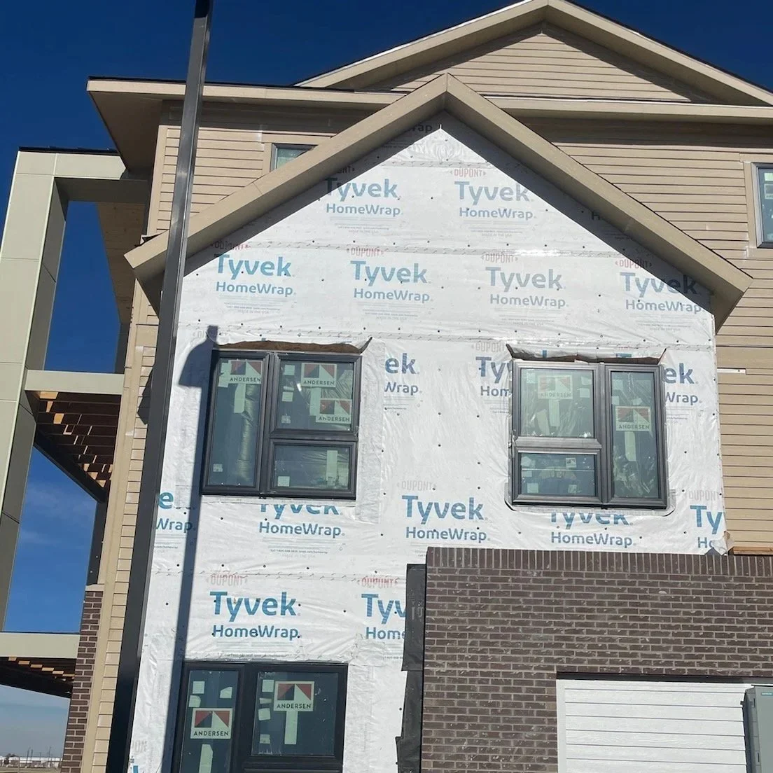A multi-story house under construction with the exterior wrapped in Tyvek HomeWrap, featuring several windows and brick accents on the lower part.