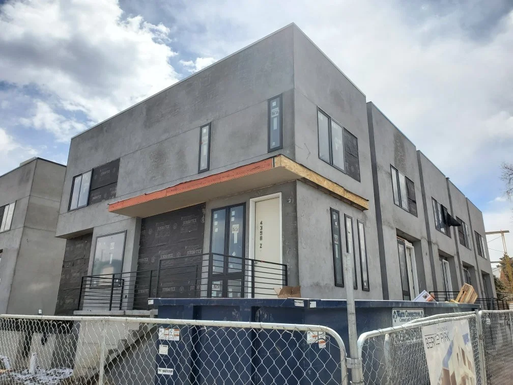 Modern multi-story residential building under construction with a gray exterior and multiple windows, surrounded by a construction fence.