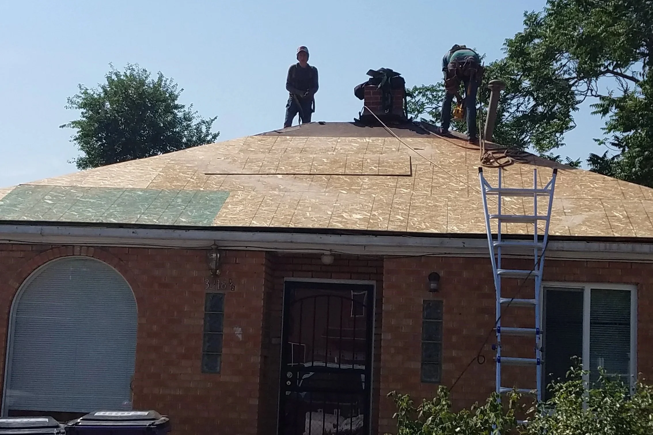 Two construction workers on the roof of a brick house, installing or repairing the shingles. A ladder is leaning against the house, and a tree is visible in the background.