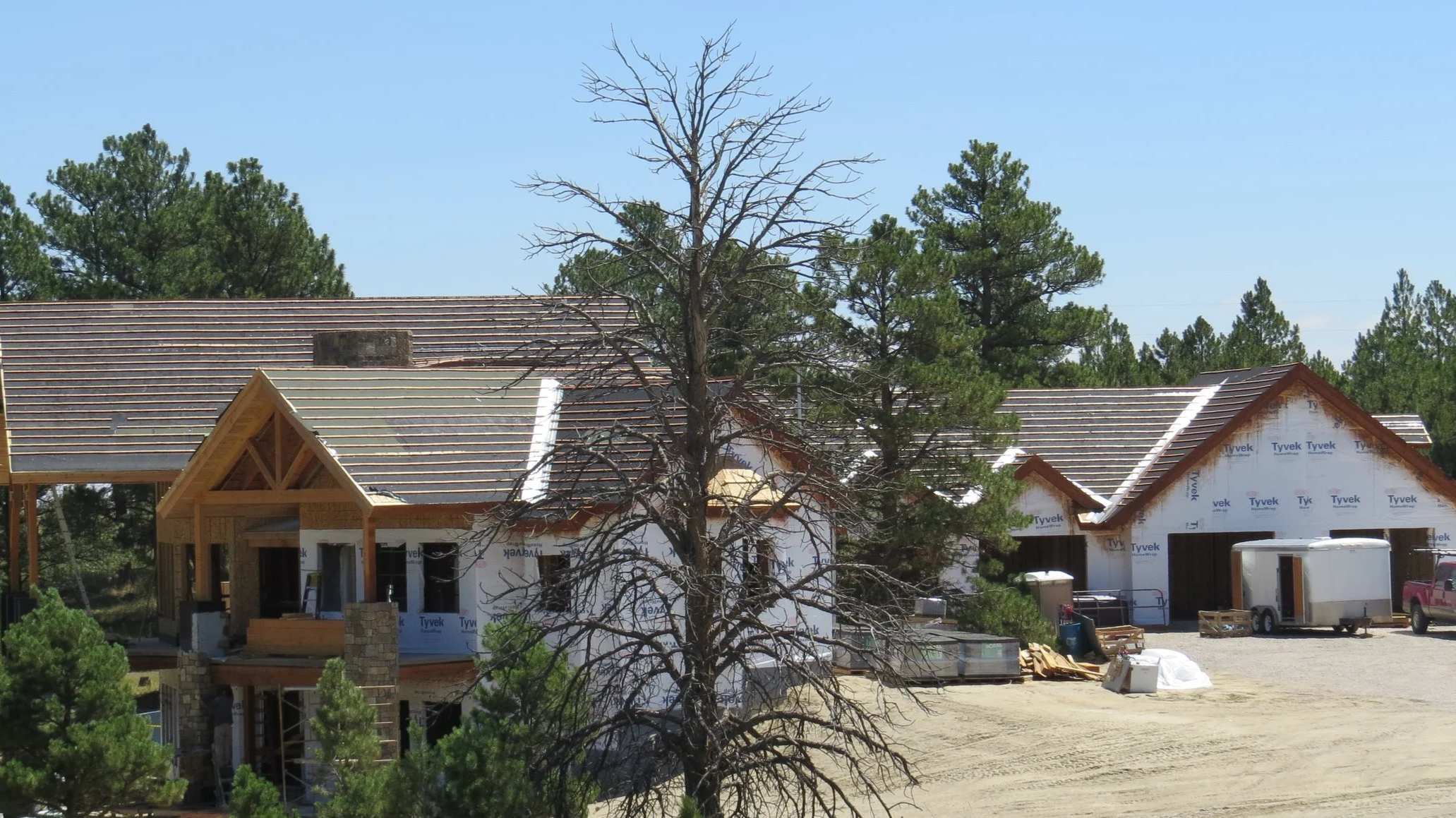 Residential construction site with houses being built, some with partially installed roofs and framing, surrounded by trees under a blue sky.